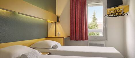 A hotel room with two beds, a TV mounted on the wall, a window with a curtain, and a wall-mounted towel rack.