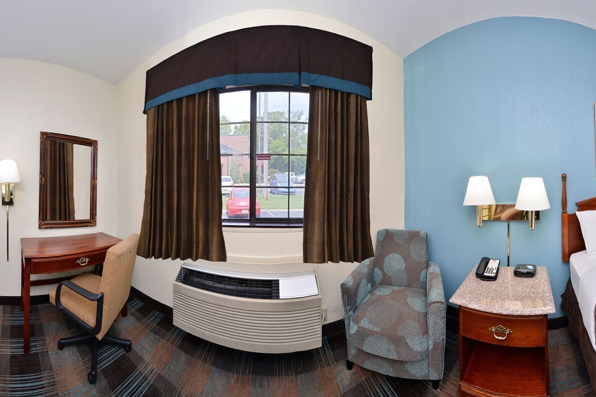 A hotel room with a bed, desk, chair, and a large window with curtains.