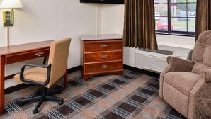 Quarto de hotel com escrivaninha, cadeira, cômoda, televisão e janela com cortinas.