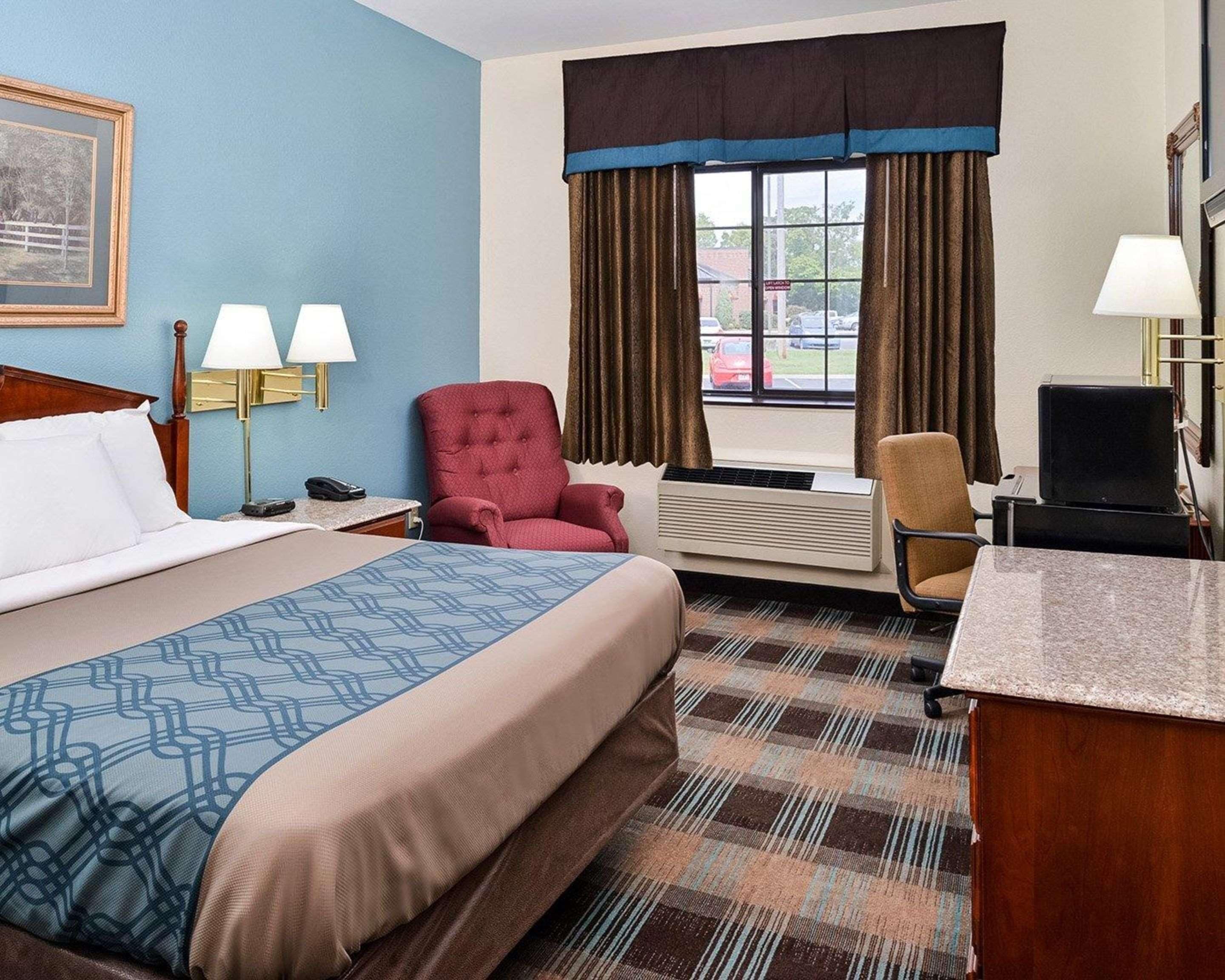 Habitación de hotel con cama, dos lámparas de noche, un sillón rojo, un escritorio con computadora y una ventana con cortinas.