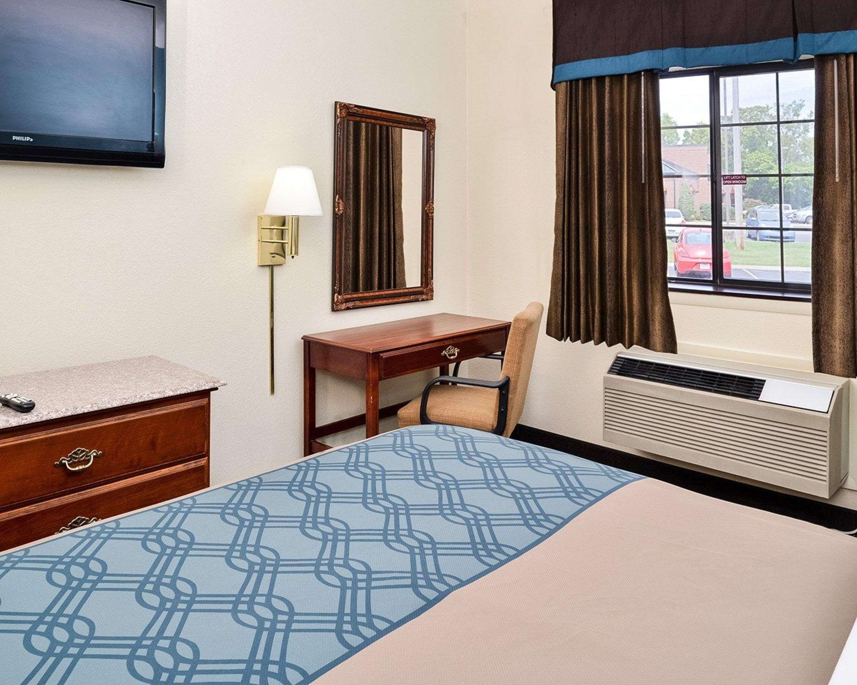 A hotel room with a bed, a desk, a chair, a television, a mirror, a lamp, and an air conditioning unit.