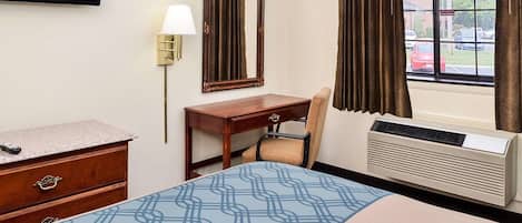 A hotel room with a bed, a desk, a chair, a television, a mirror, a lamp, and an air conditioning unit.