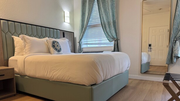 A neatly made bed with a headboard, a bedside table, and a window with curtains. - Athens Hotel Suites (Houston)