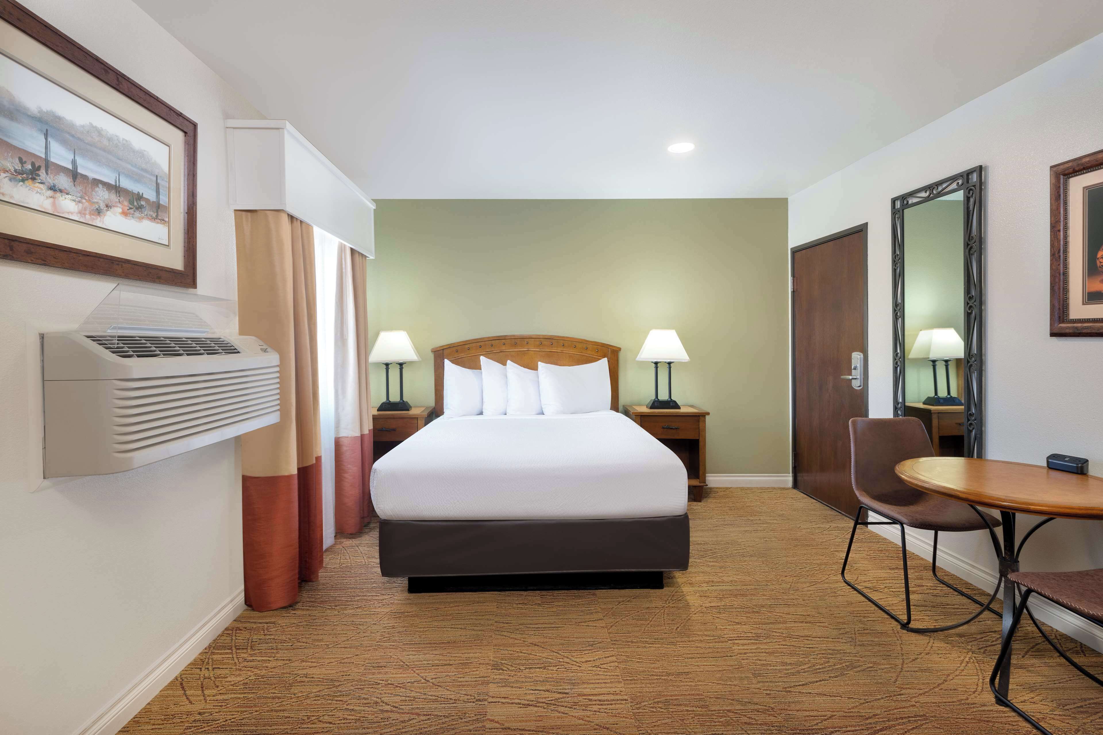Quarto de hotel com uma cama, duas mesas de cabeceira com luminárias, uma escrivaninha com cadeira, um espelho grande e ar-condicionado fixado na parede.