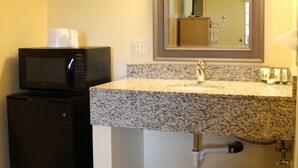 A compact bathroom with a granite countertop sink, a mirror above it, a microwave, and a trash can. - BeachWay Inn (Arroyo Grande)