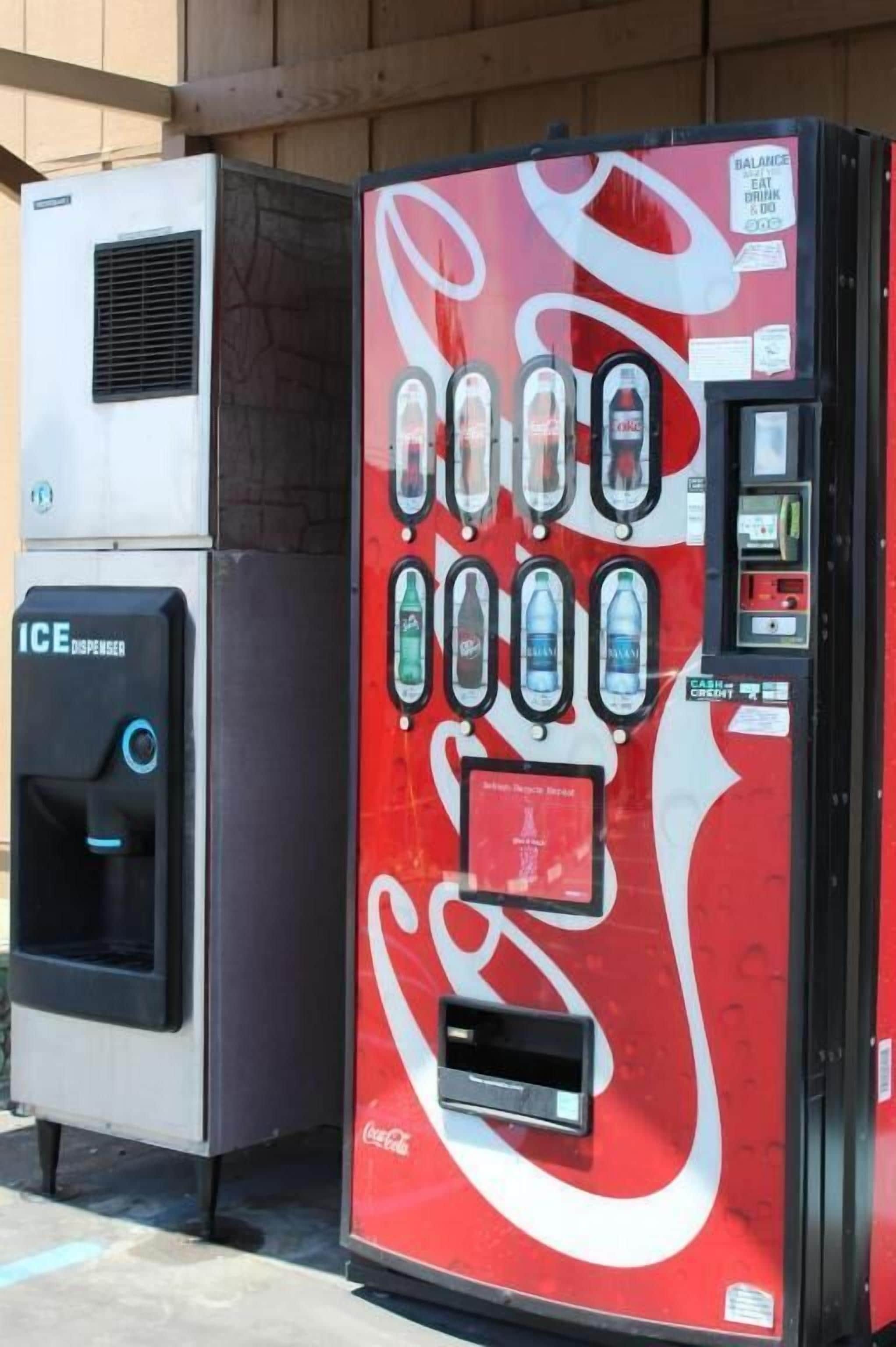 Eine Coca-Cola-Getränkeautomaten mit Coca-Cola-Logo, einem Bargeldautomaten und einem Schild mit der Aufschrift 'BALANCE EAT DRINK & DO'.