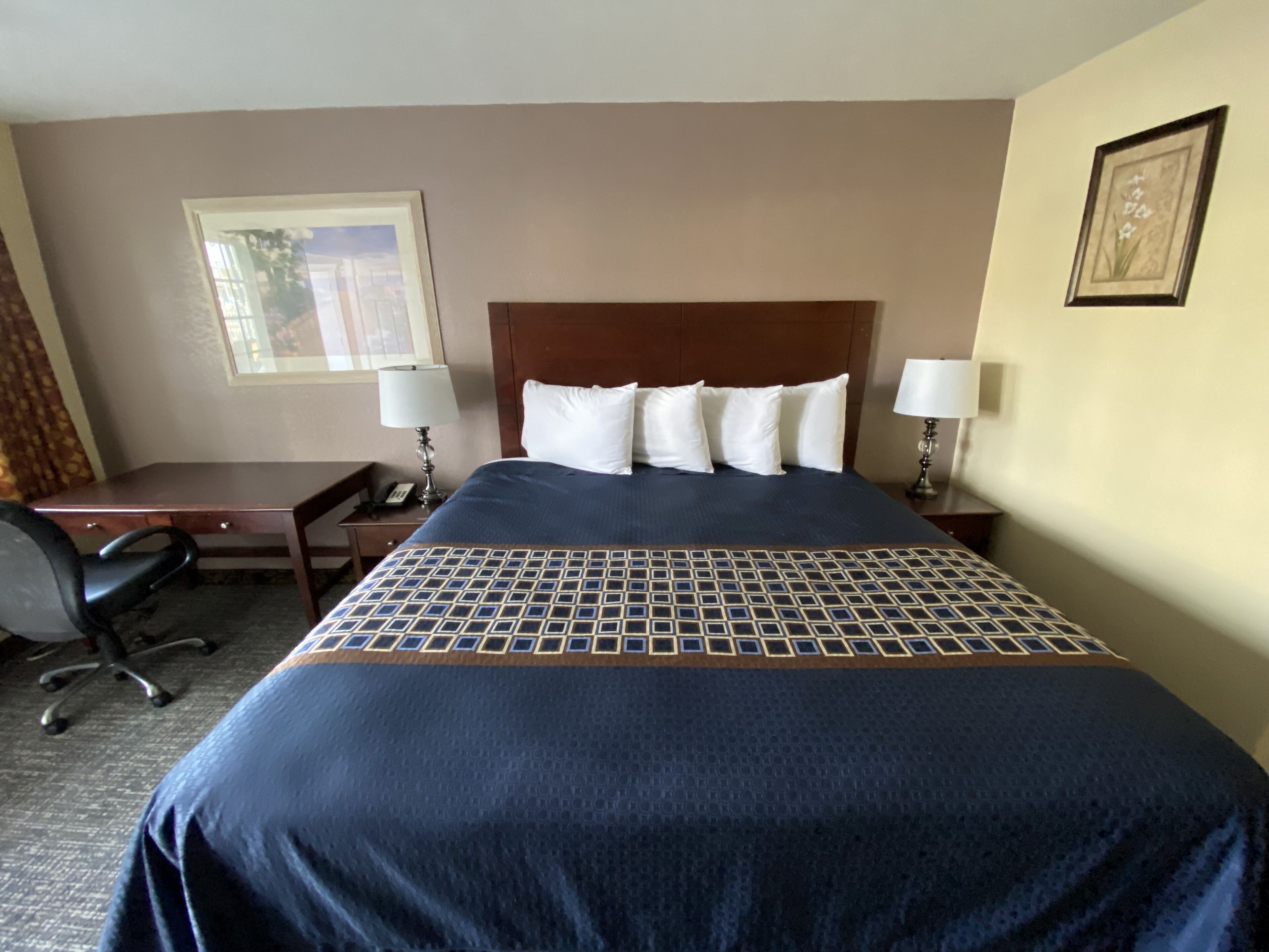 A hotel room with a large bed, a desk, two chairs, and a nightstand with a lamp.