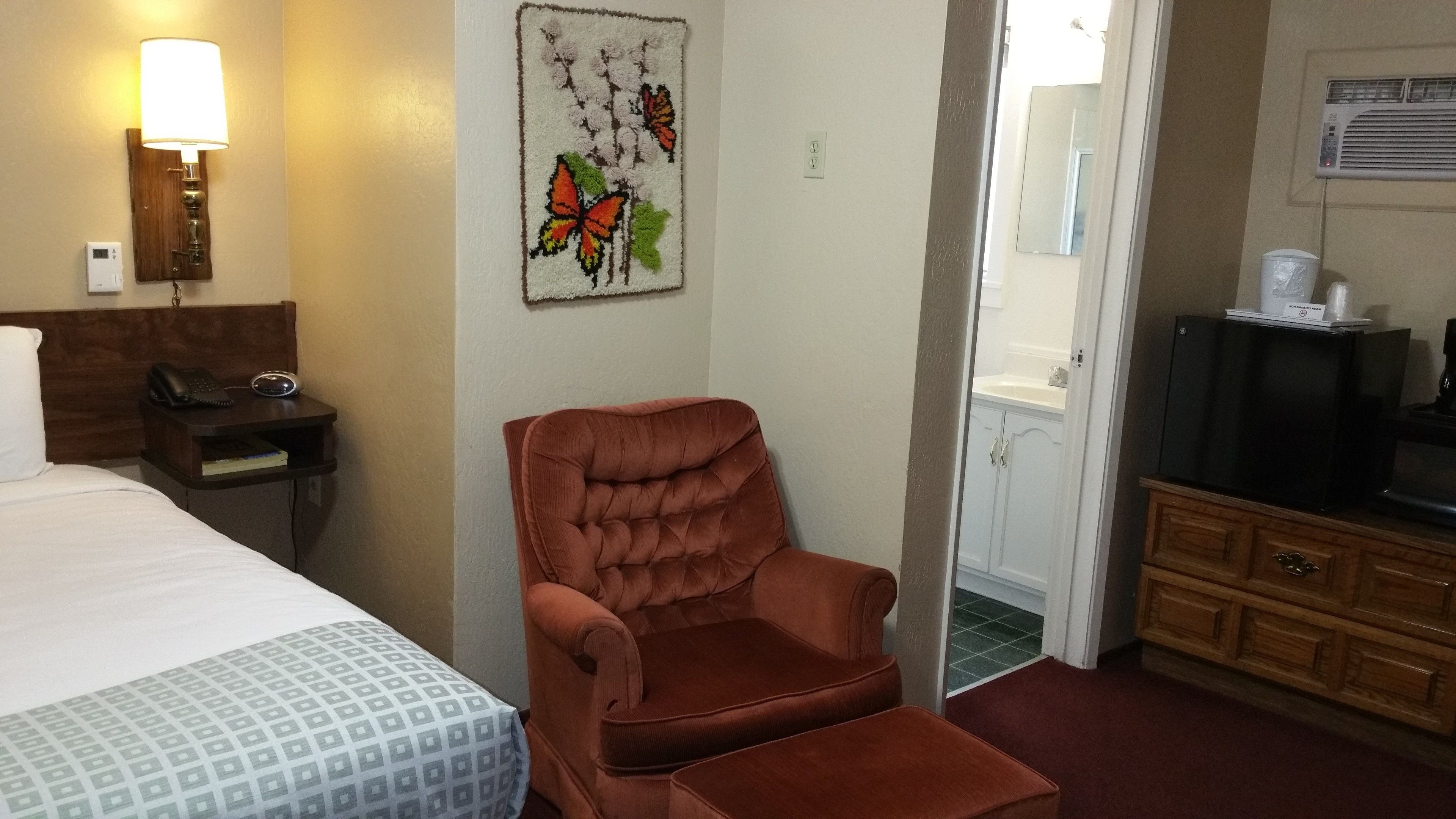 A hotel room with a bed, a brown armchair, a desk with a phone and a lamp, a painting of butterflies, and a TV on a cabinet.