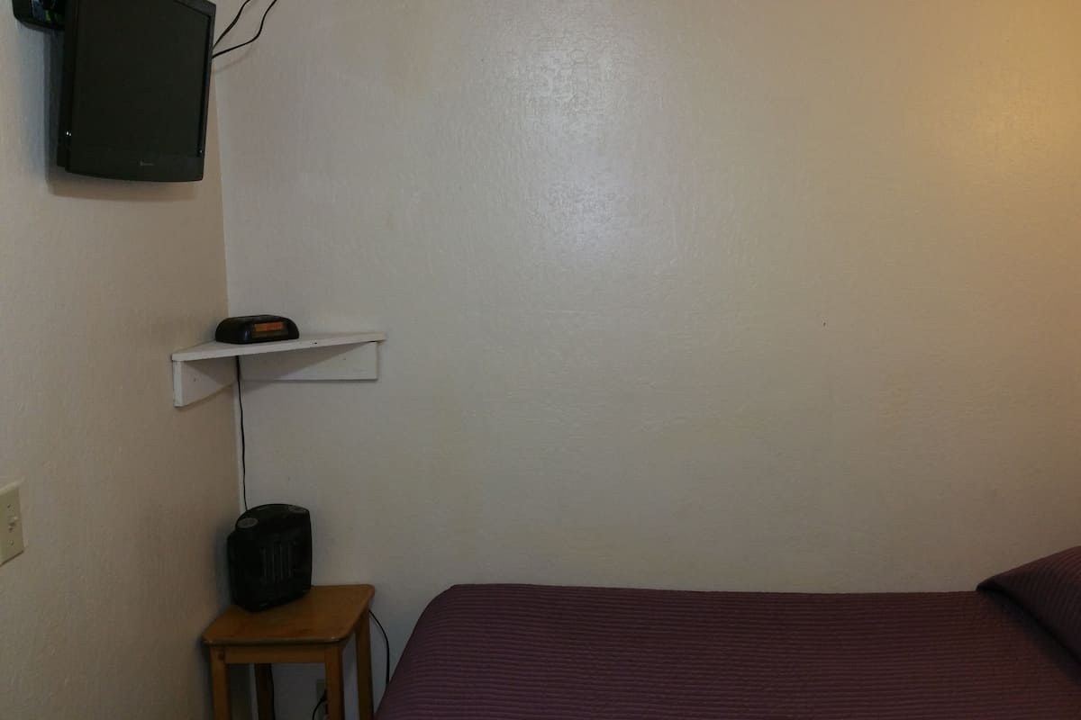A small, simple room with a bed, a small table, a shelf, and a speaker.