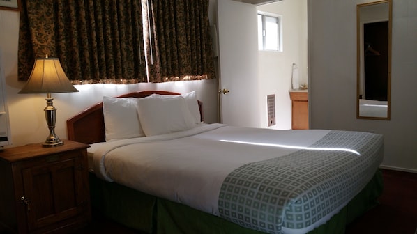 Una cama bien tendida con sábanas blancas y un plaid estampado, una mesita de noche de madera con una lámpara, y una ventana con cortinas.