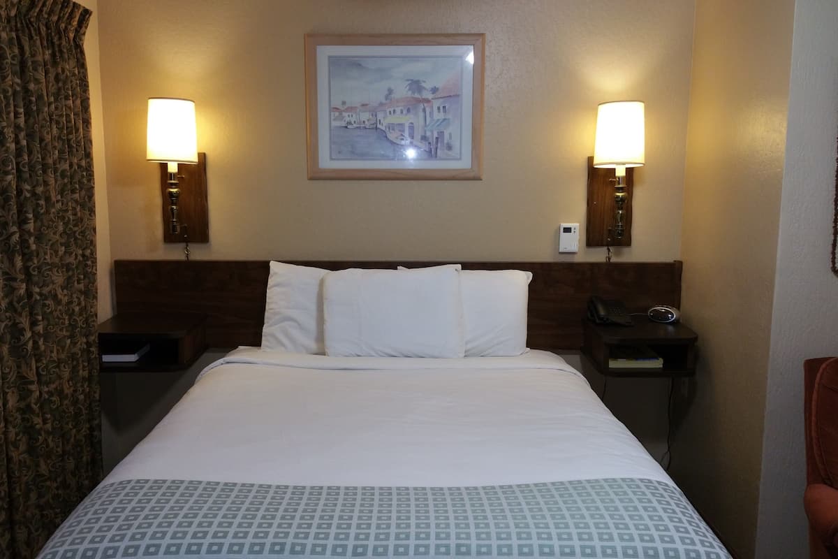 A hotel room with a bed, two bedside lamps, a nightstand with a phone, a wall clock, and a framed picture above the bed.