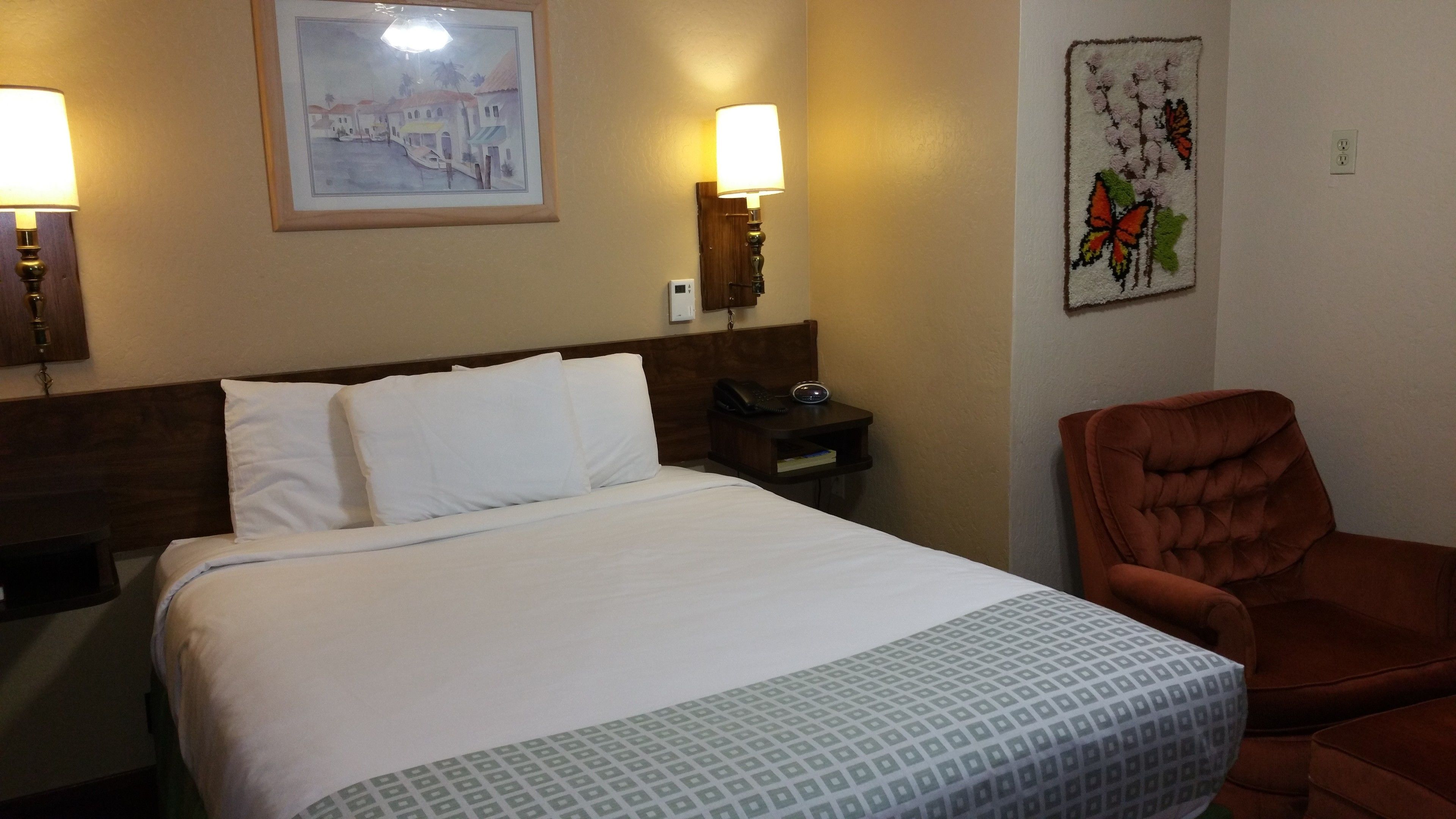 A hotel room with a bed, two bedside lamps, a nightstand with a phone, a chair, and a wall with a framed picture and a tapestry.