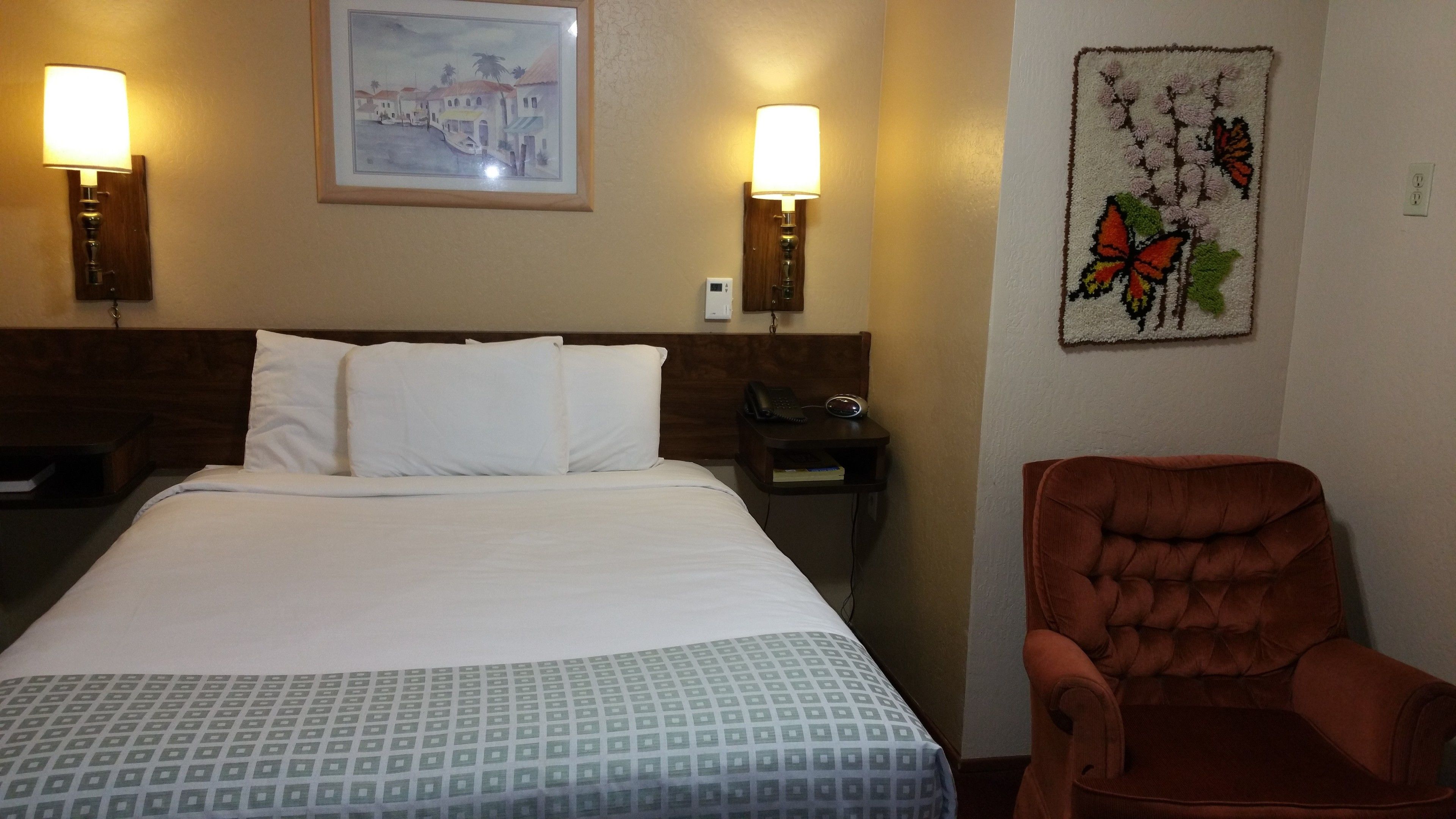 A hotel room with a large bed, two bedside lamps, a framed painting, a decorative wall hanging, and a brown armchair.