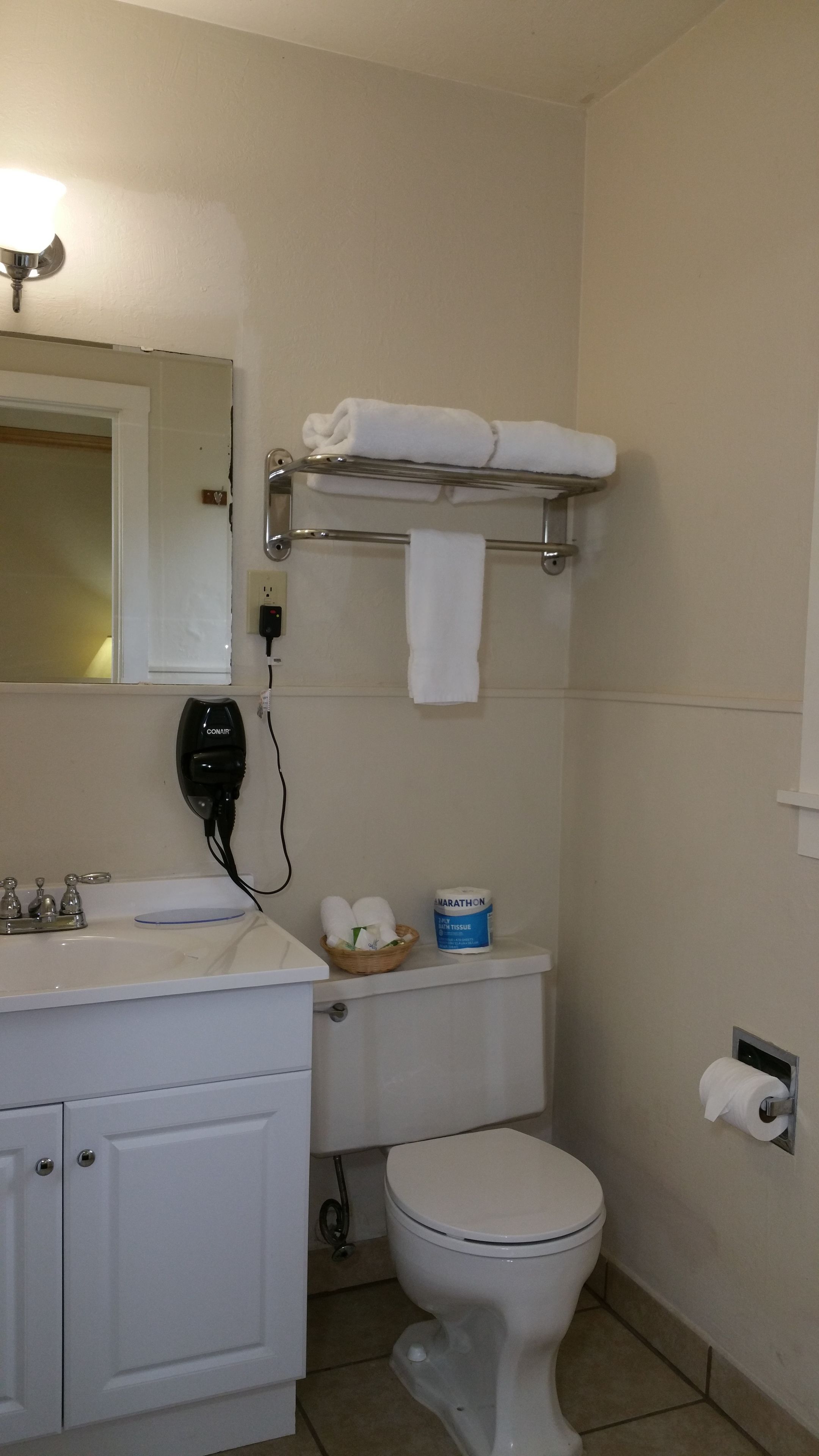 Un baño con lavamanos blanco, espejo, toallero con toallas, inodoro y dispensador de papel higiénico.