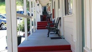 A covered porch with red steps leading to a door, outdoor seating, and a view of parked cars. - El Dorado Motel (Twain Harte)