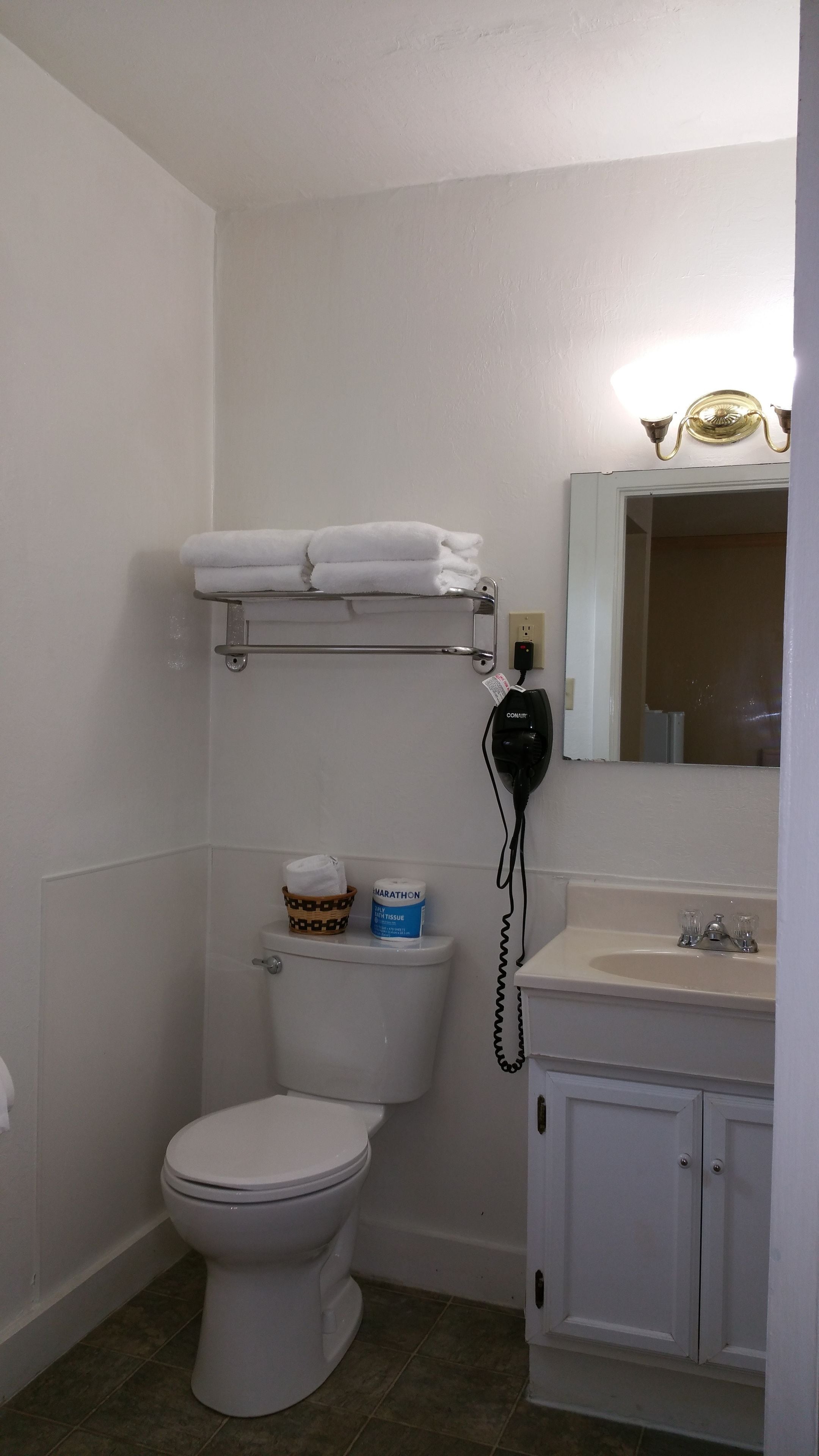 Un baño con inodoro, un estante con toallas, dispensador de papel higiénico, un cubo de basura y un lavamanos con espejo arriba.