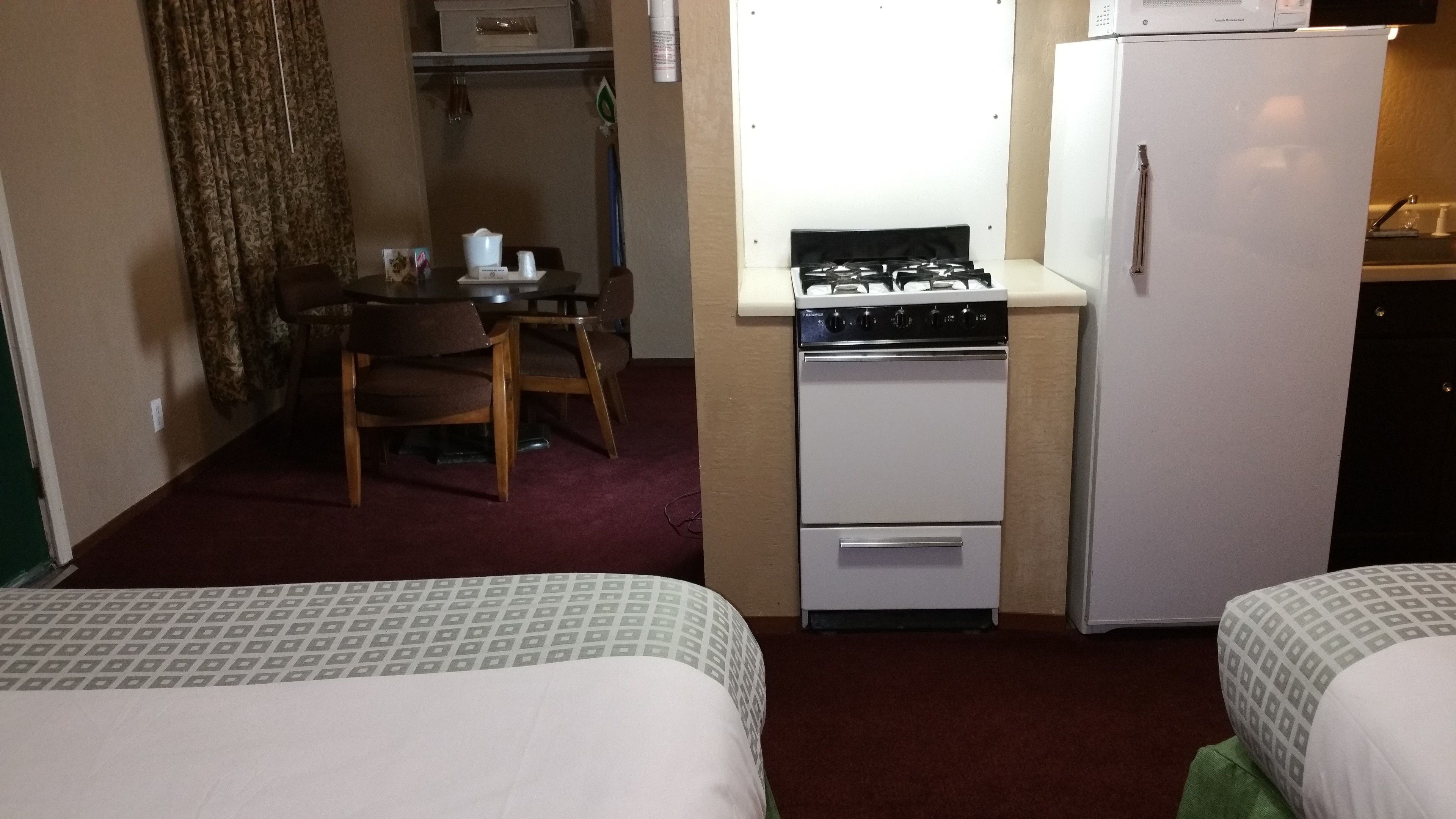 Quarto de hotel compacto com duas camas, pequena cozinha equipada com fogão e geladeira, mesa de jantar com cadeiras e piso coberto por carpete.