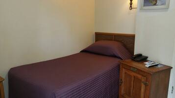 Un lit simple avec une tête de lit en bois et une couvre-lit violette. Il y a une table de chevet en bois sur laquelle se trouvent un téléphone et une carte.