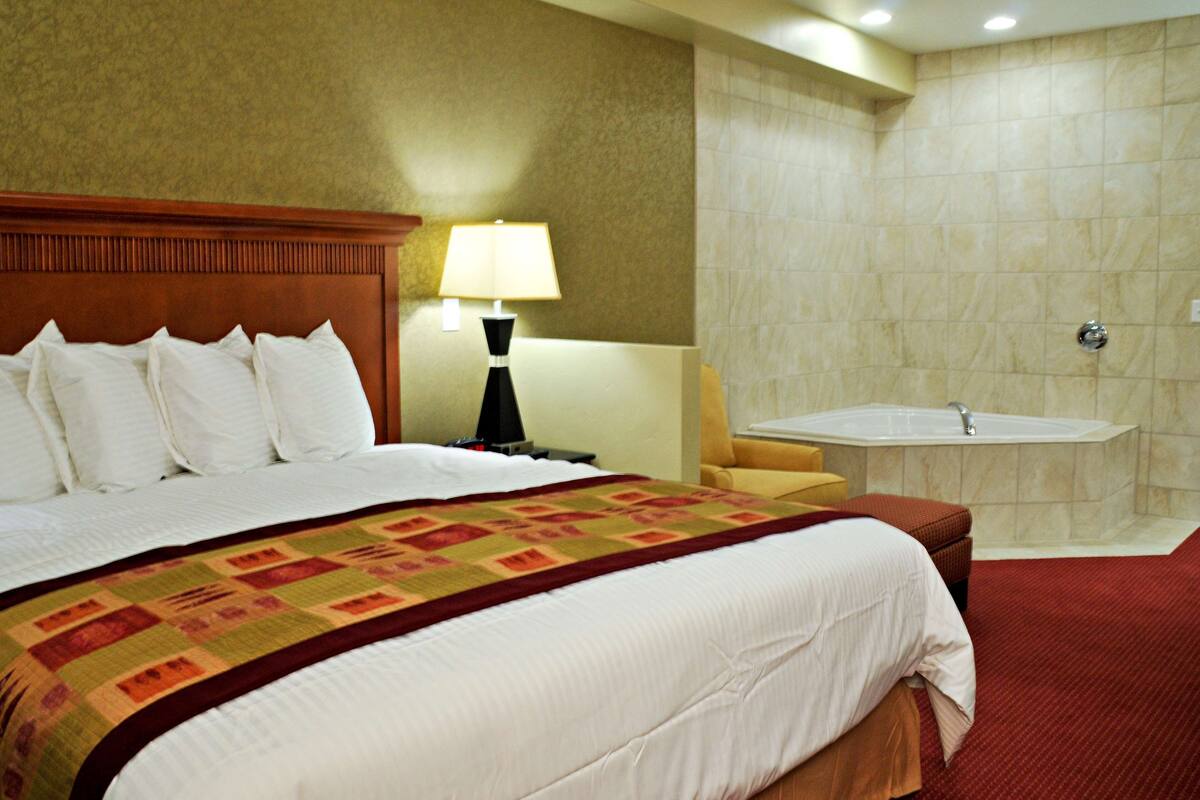 A hotel room with a large bed, a nightstand with a lamp, a bathroom with a bathtub, and a red carpet.