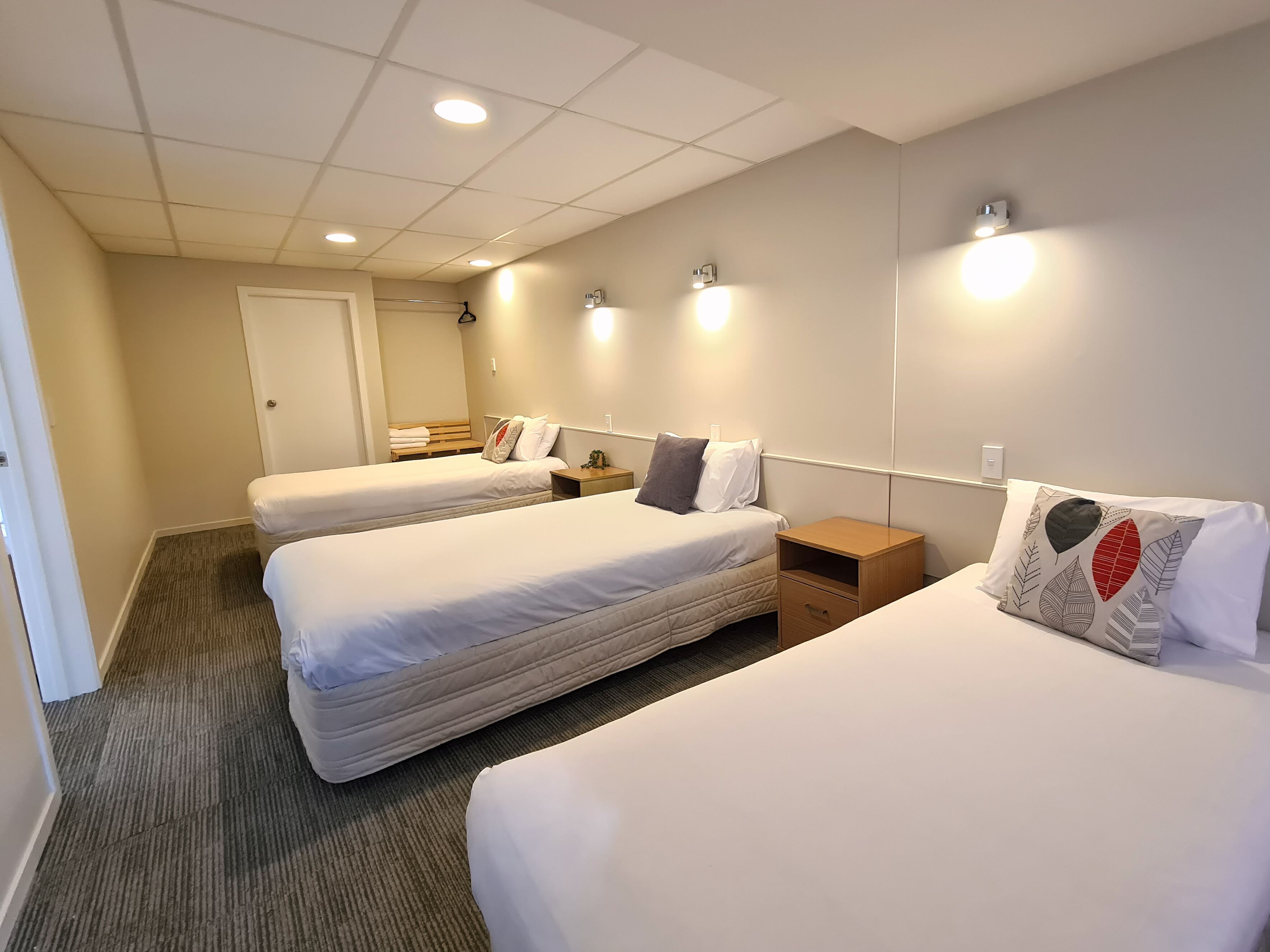 Ett hotellrum med tre enkelsängar, ett litet träbord vid sängen, en liten träbyrå och ett golv täckt med matta.
