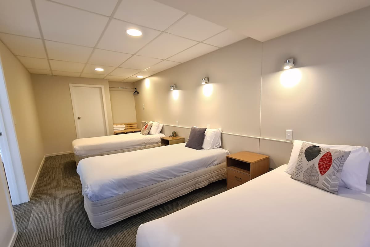 Une chambre d’hôtel avec trois lits simples, une table de chevet en bois, une petite commode en bois et un sol recouvert de moquette.