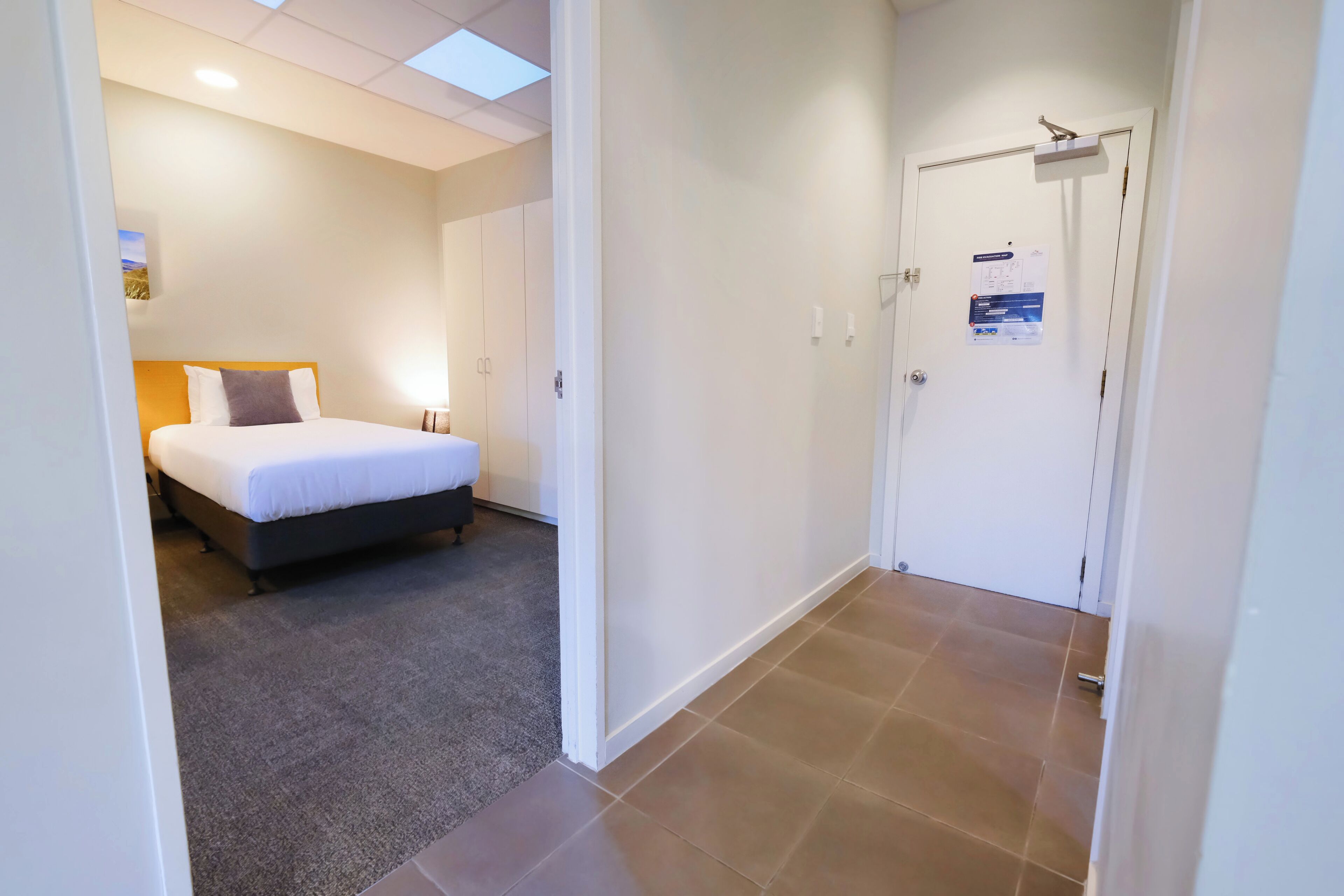 A hotel room with a bed, a carpeted floor, a white door, and a light switch on the wall.