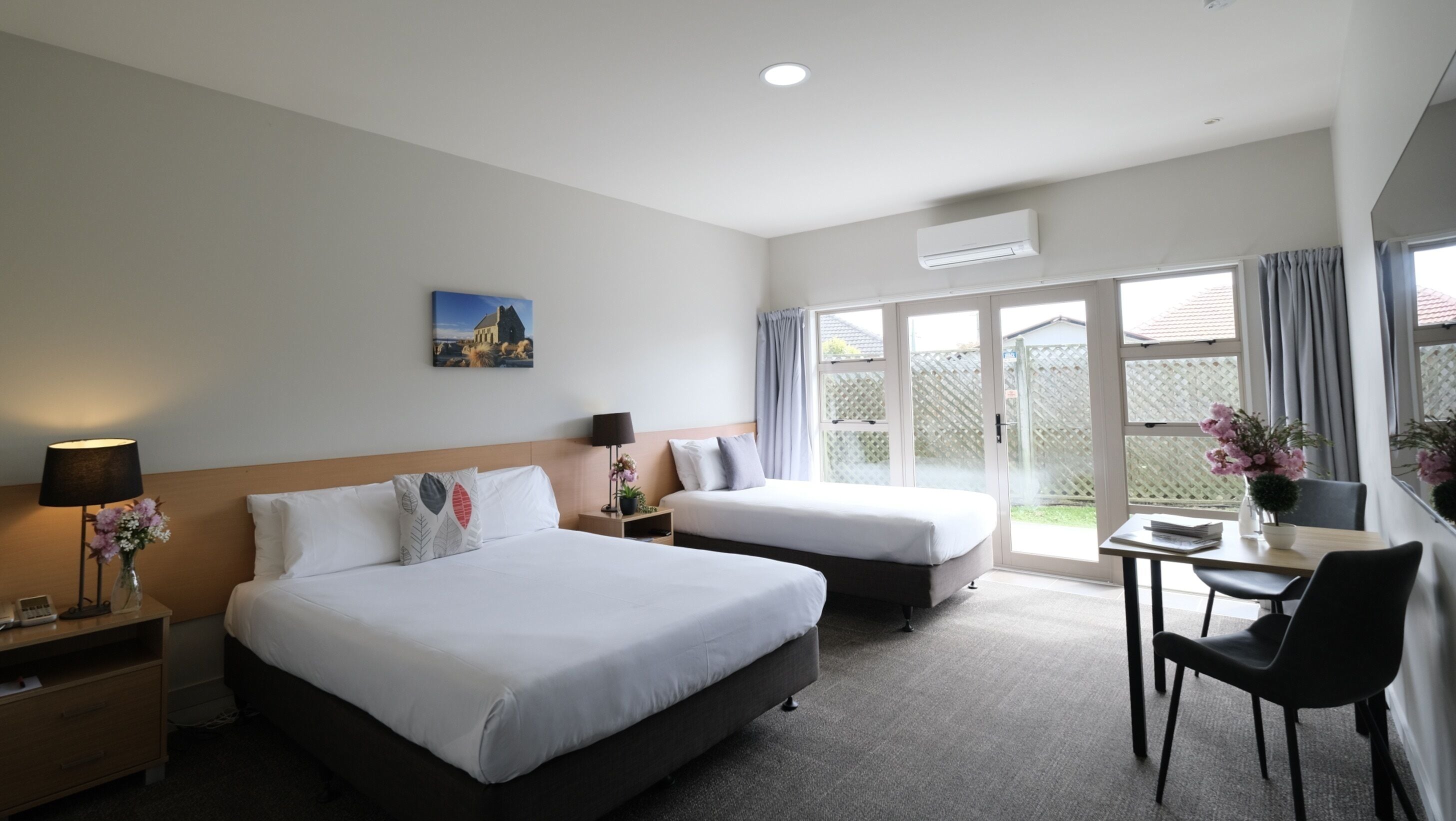 Habitación de hotel con dos camas, un escritorio, una silla, una lámpara, un jarrón con flores y un cuadro en la pared.