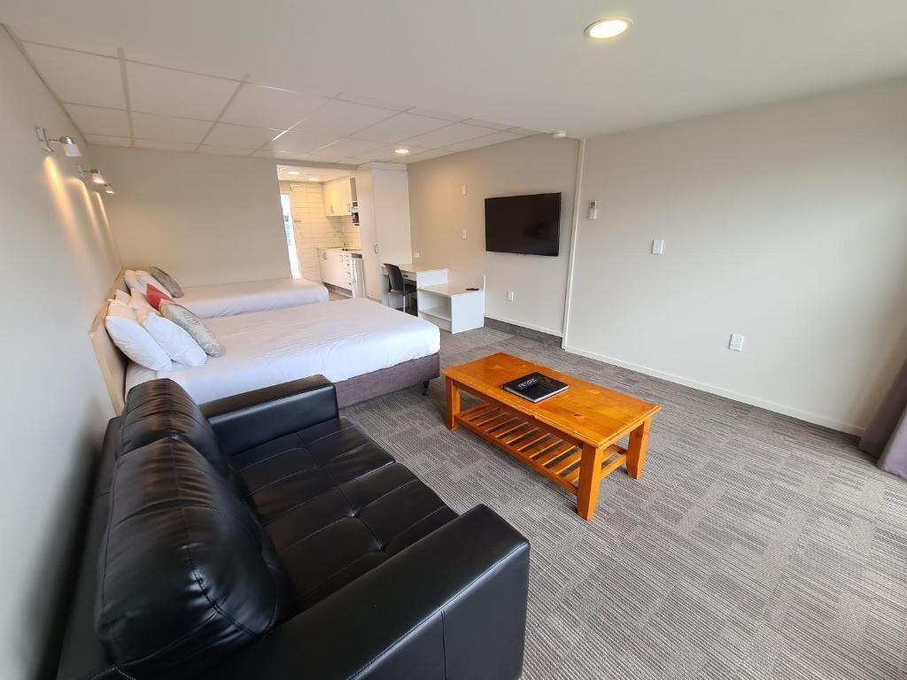 A modern hotel room with a bed, a black leather sofa, a wooden coffee table, and a flat-screen TV mounted on the wall.