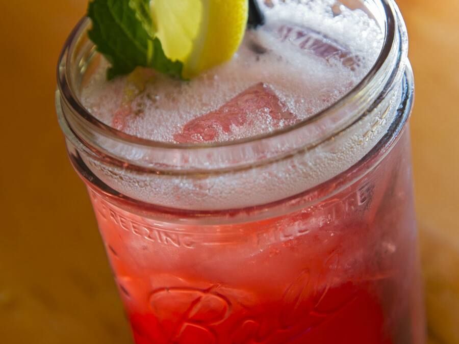 A glass jar with a red beverage, ice cubes, and a lemon slice with mint leaves.