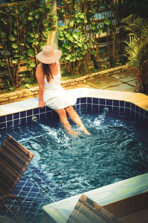 Una persona vestida de blanco sentada en el borde de un jacuzzi pequeño y azulejado, rodeado de follaje.