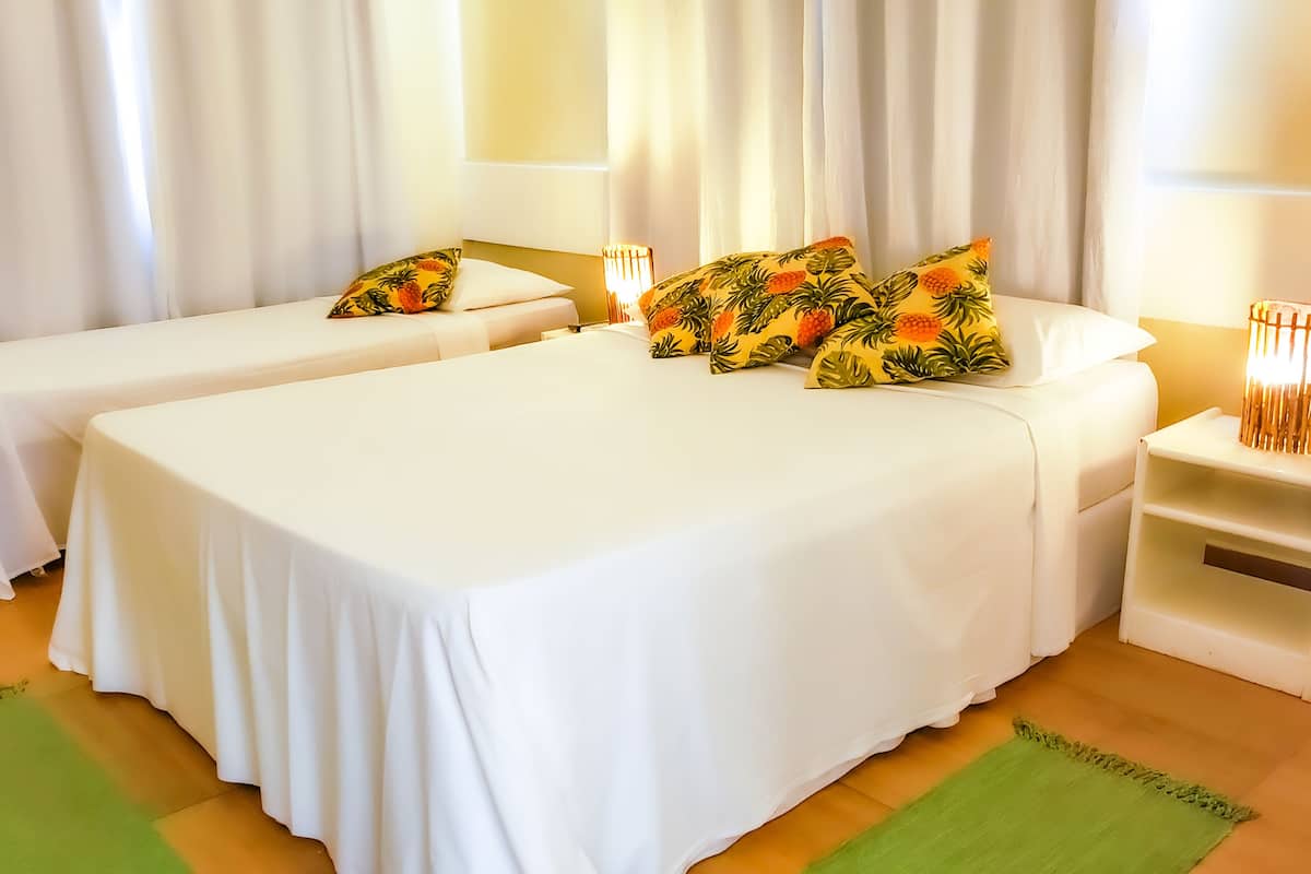 Habitación de hotel con dos camas individuales, ropa de cama blanca y almohadas con motivos tropicales. Hay mesitas de noche con lámparas y un pequeño estante.
