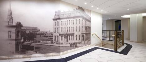 Un pasillo con un gran mural de un edificio histórico, una escalera con barandilla de metal y piso de baldosas.