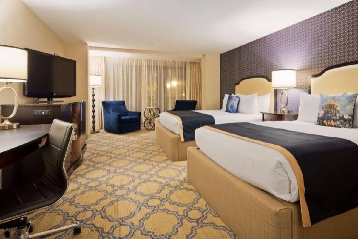 Une chambre d’hôtel avec deux lits, un bureau avec une télévision, une chaise et une moquette à motifs.