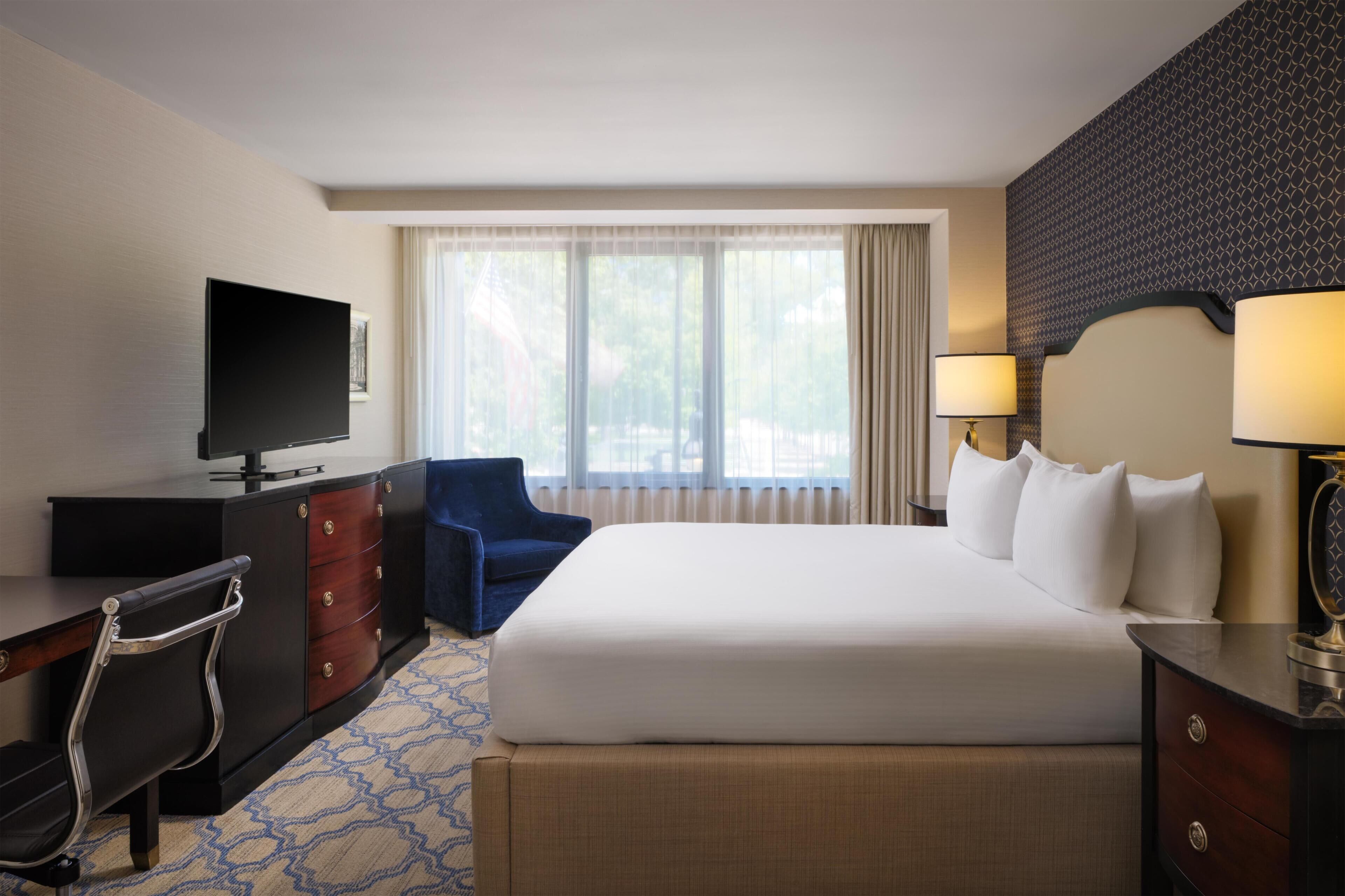 A hotel room with a bed, a desk with a TV, a chair, a dresser, and a window with curtains.