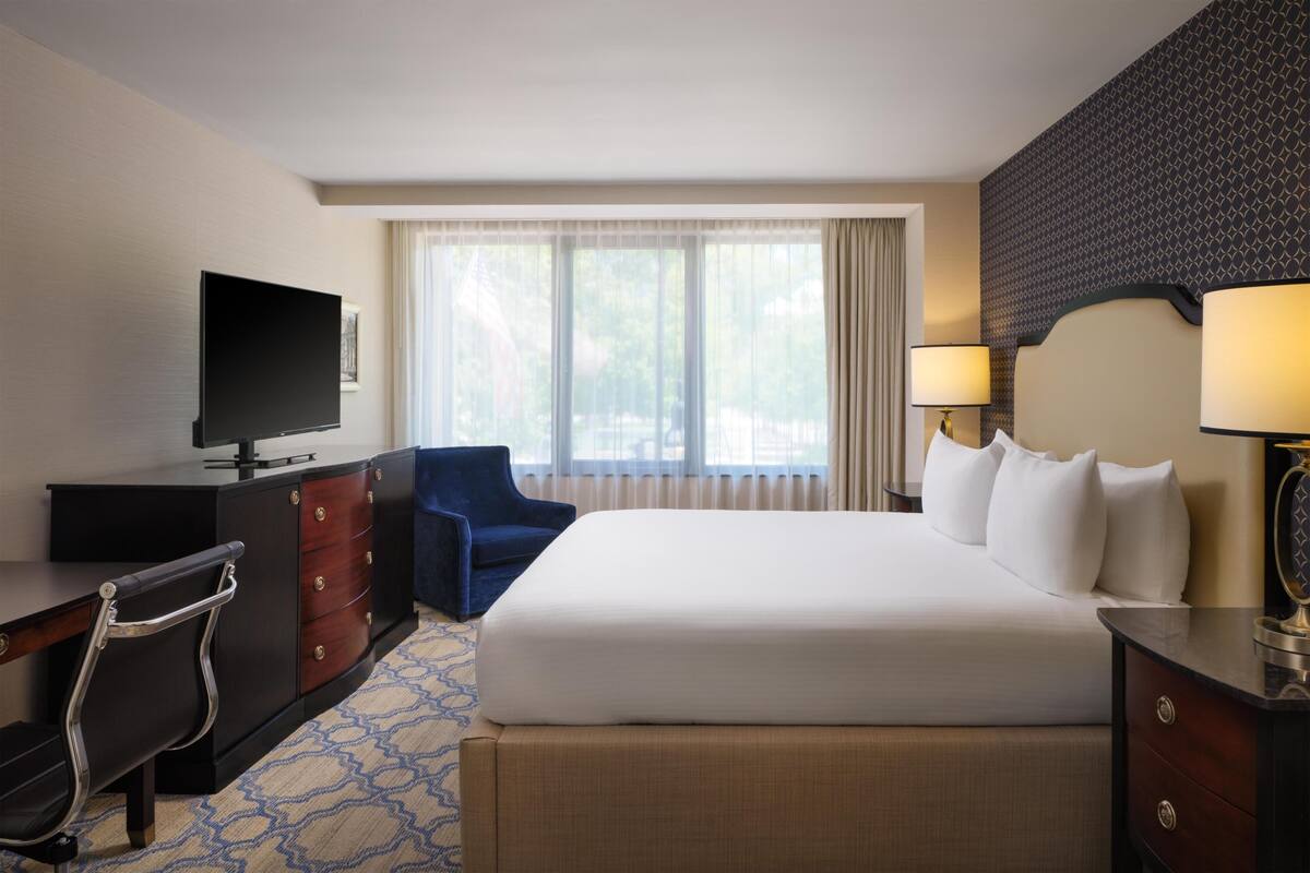 Une chambre d’hôtel avec un lit, un bureau avec une télévision, une chaise, une commode et une fenêtre avec des rideaux.