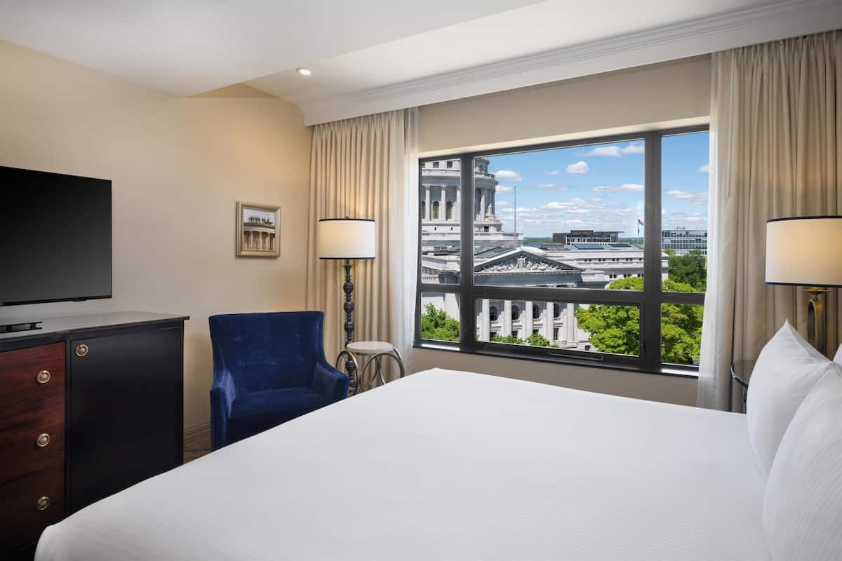 Une chambre d’hôtel avec un lit, une télévision, une chaise bleue, une petite table et une grande fenêtre donnant sur un immeuble.