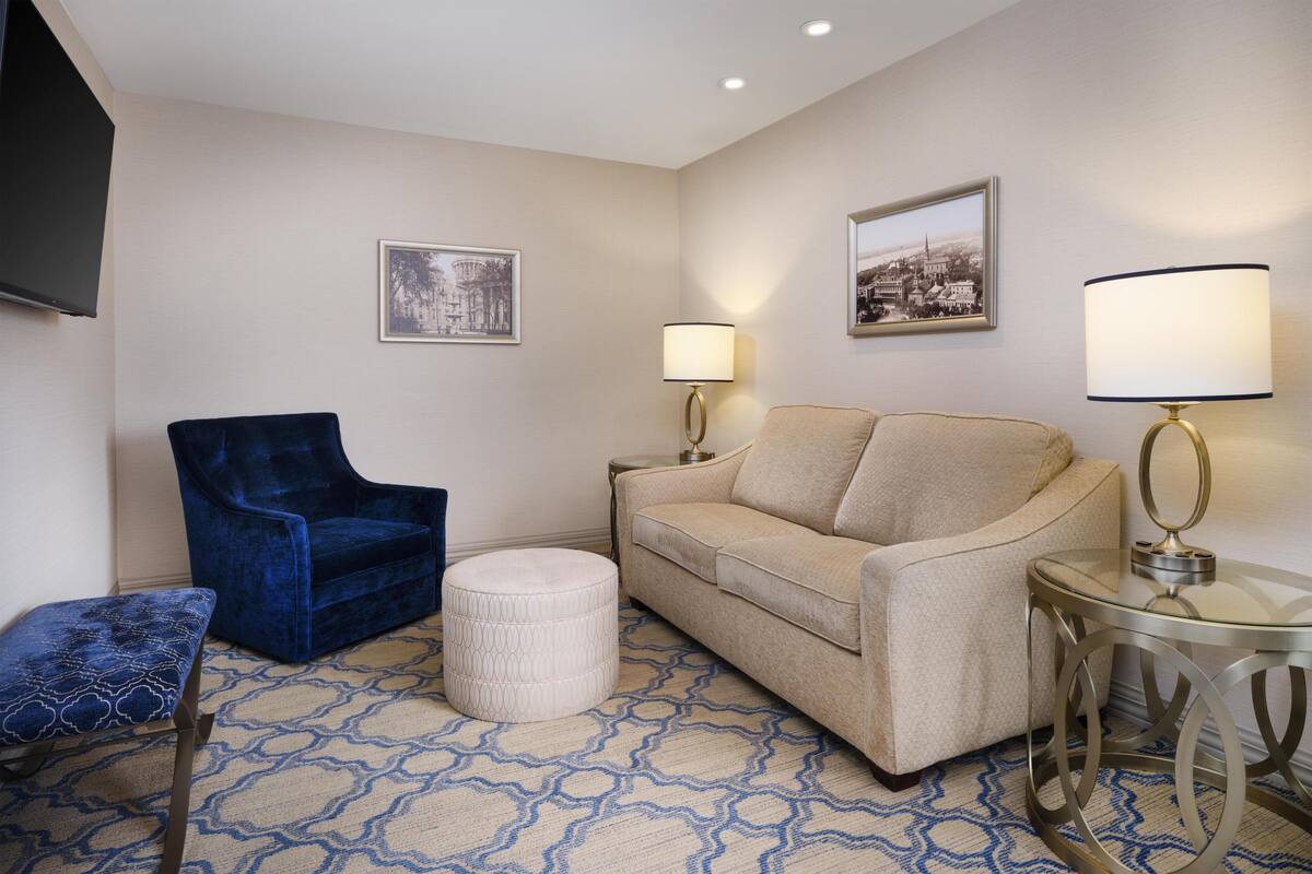 Une pièce comprenant un fauteuil bleu, un canapé beige, un pouf rond, une table en verre et deux lampes fixées au mur.