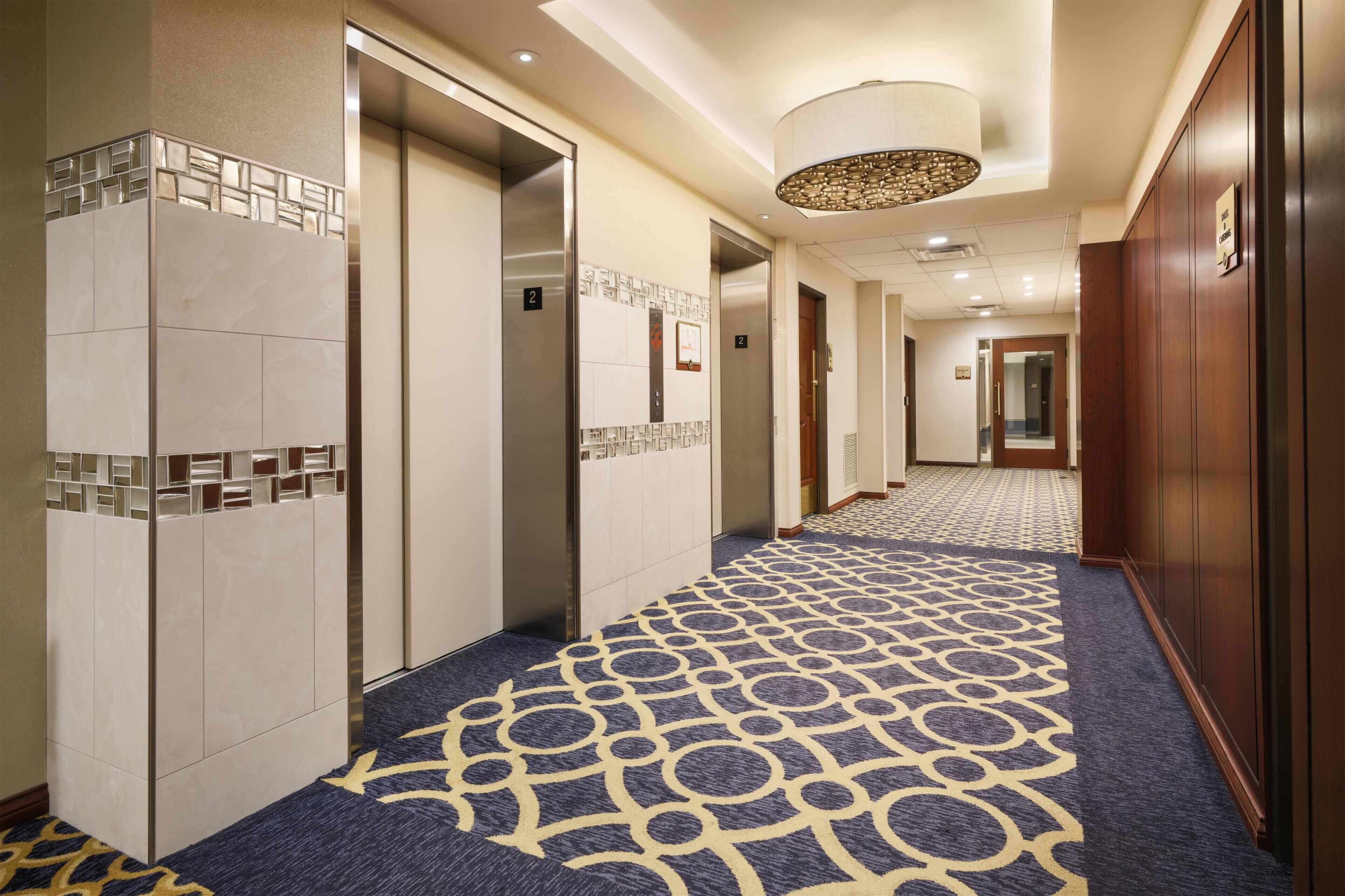 Un couloir d’hôtel avec des ascenseurs, une moquette à motifs et un luminaire moderne.