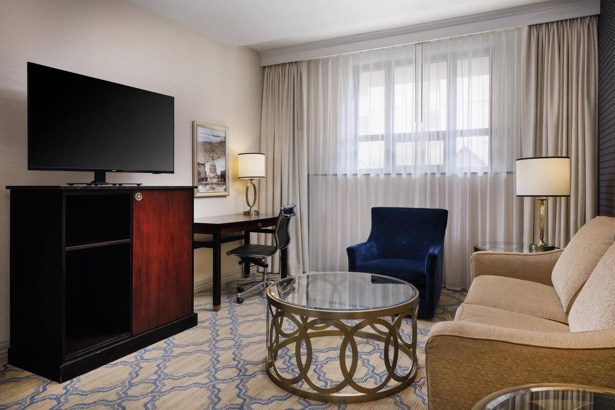 Une chambre d’hôtel équipée d’un téléviseur à écran plat, d’une table basse en verre, d’un canapé, d’un fauteuil, d’un bureau et d’une lampe.