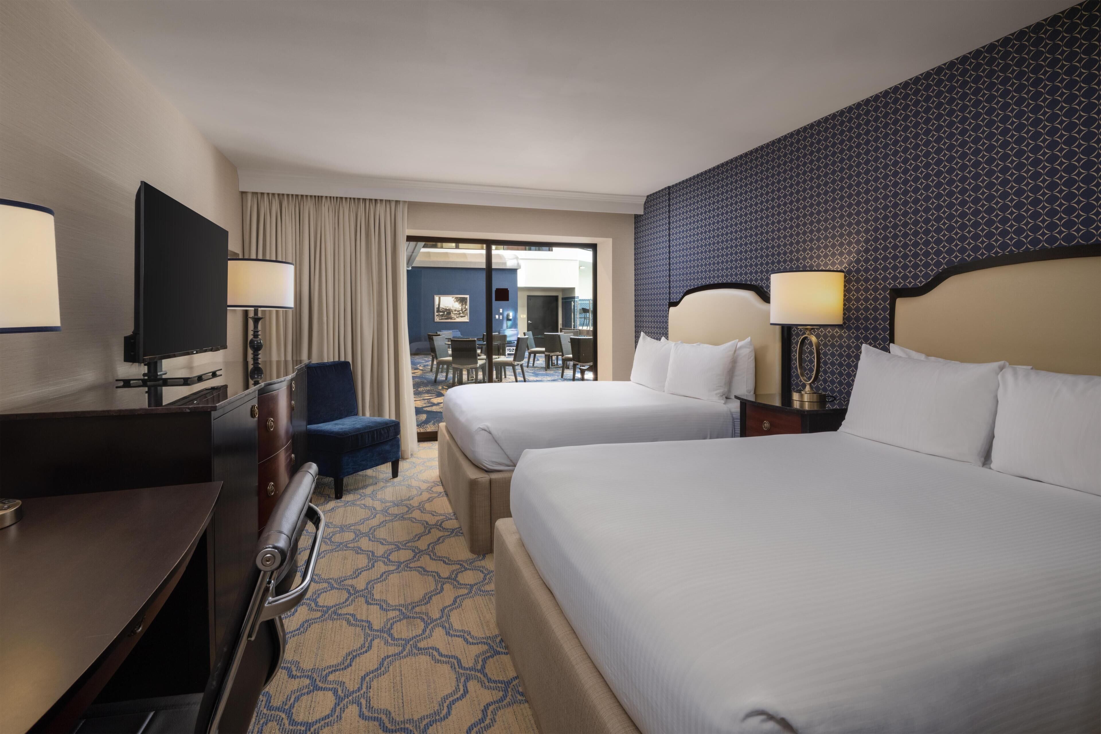 A hotel room with two beds, a desk with a television, a chair, a lamp, and a view of the outdoor seating area.