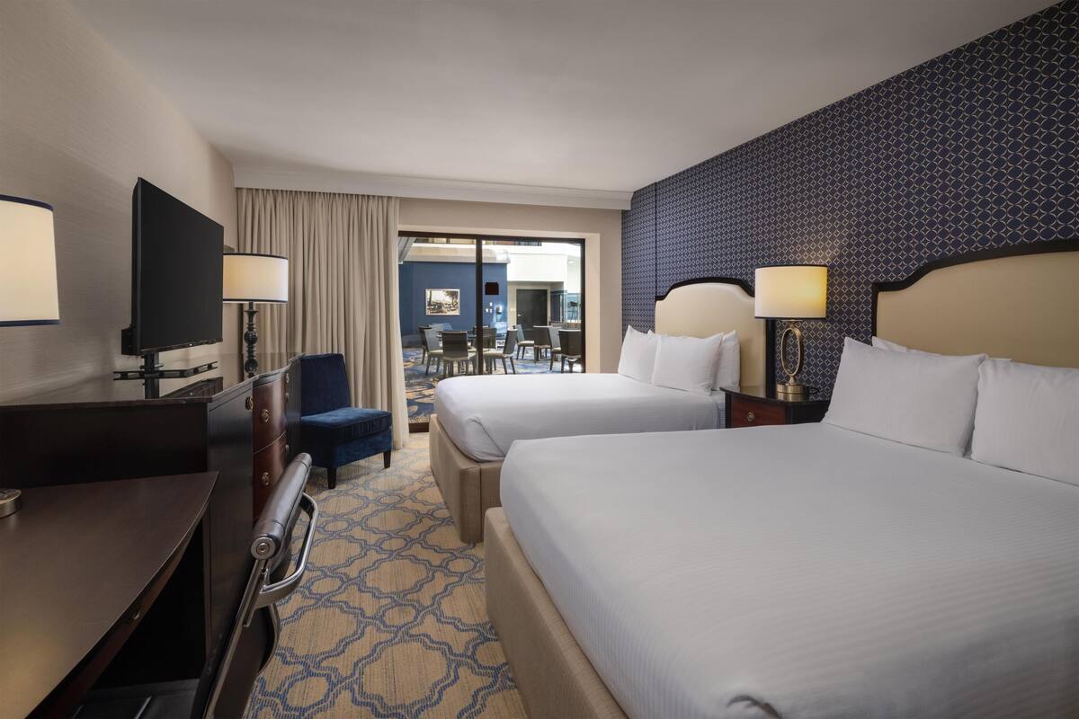 Une chambre d’hôtel avec deux lits, un bureau avec une télévision, une chaise, une lampe et une vue sur l’espace extérieur aménagé en coin salon.
