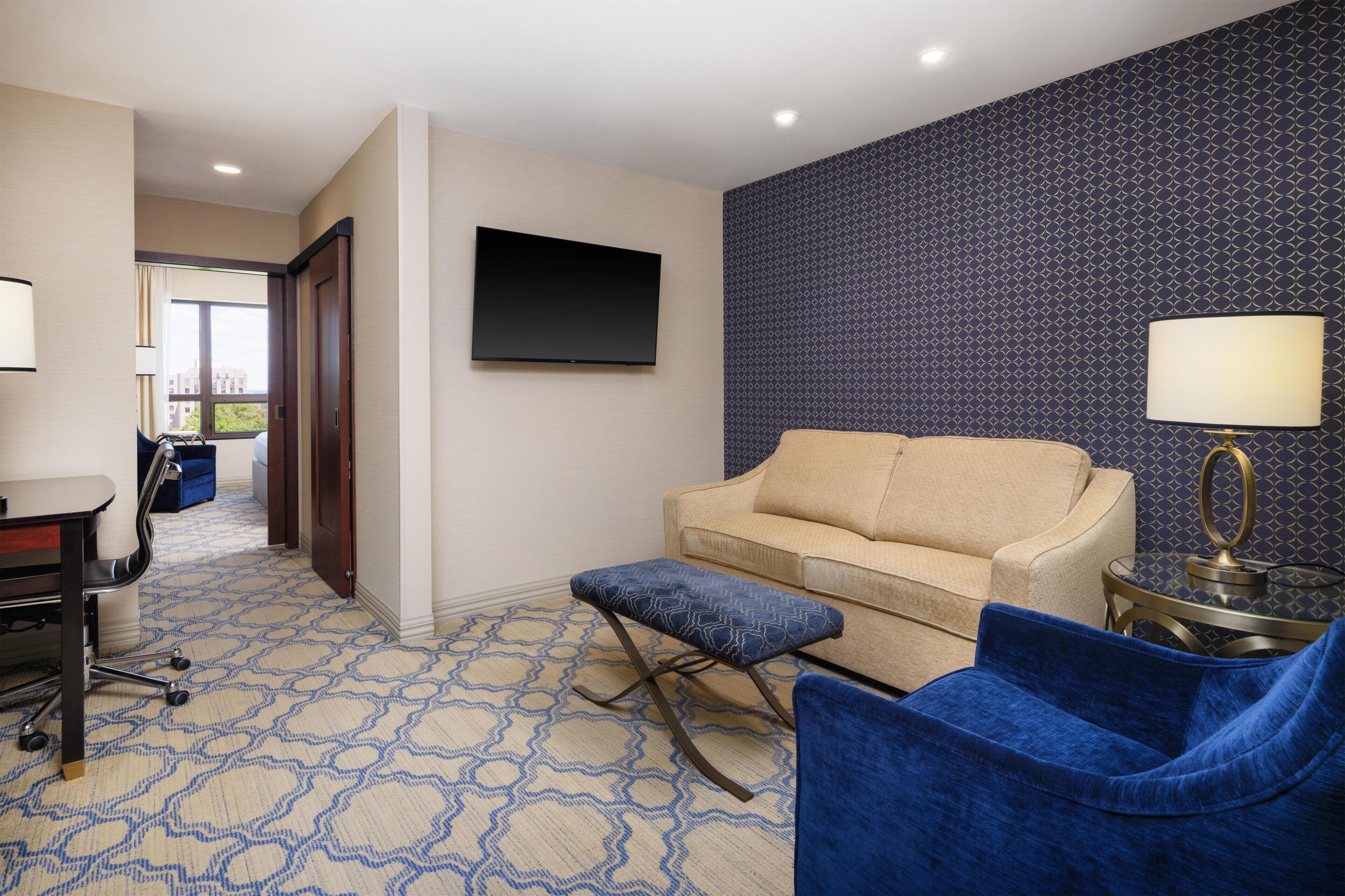 A hotel room with a sofa, ottoman, blue chair, TV, and a patterned carpet.