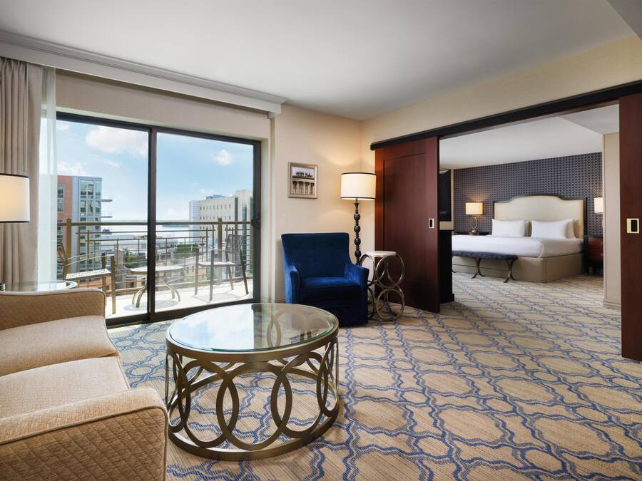 Ein Hotelzimmer mit Balkon, einem Bett, einem blauen Sessel, einem runden Glastisch und einem gemusterten Teppich.