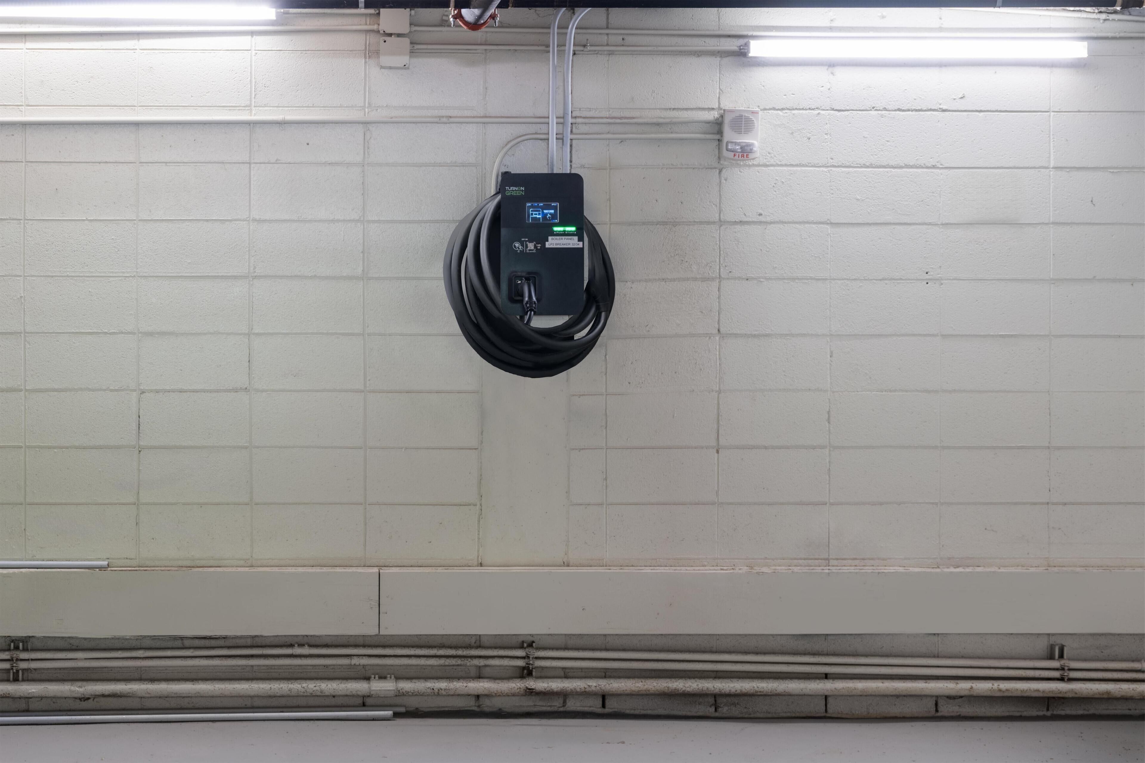 Una caixa de subministraments amb una llum indicadora verda, un cable enrotllat i un detectador de foc a una paret amb rajoles blanques.