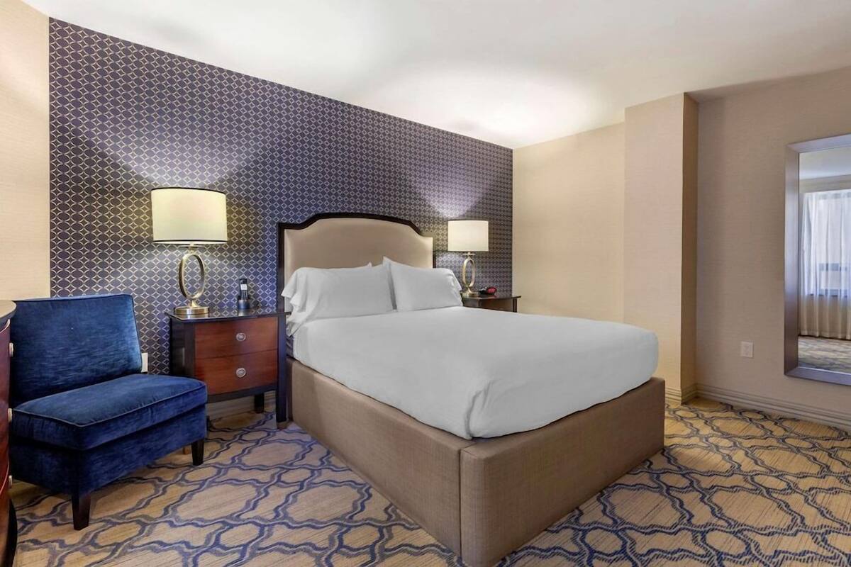 Une chambre d’hôtel avec un lit, deux tables de chevet avec des lampes, une chaise bleue, une moquette à motifs et une fenêtre avec des rideaux.