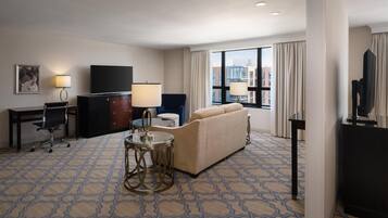 A hotel room with a sofa, TV, desk, lamp, and a view of the city.