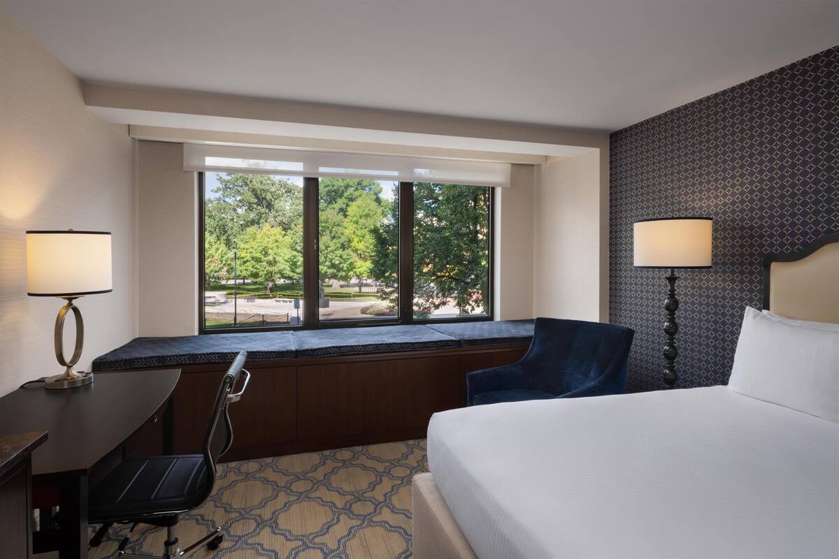 Une chambre d’hôtel avec un lit, un bureau, une chaise, une fenêtre donnant sur des arbres et un mur décoré de motifs.
