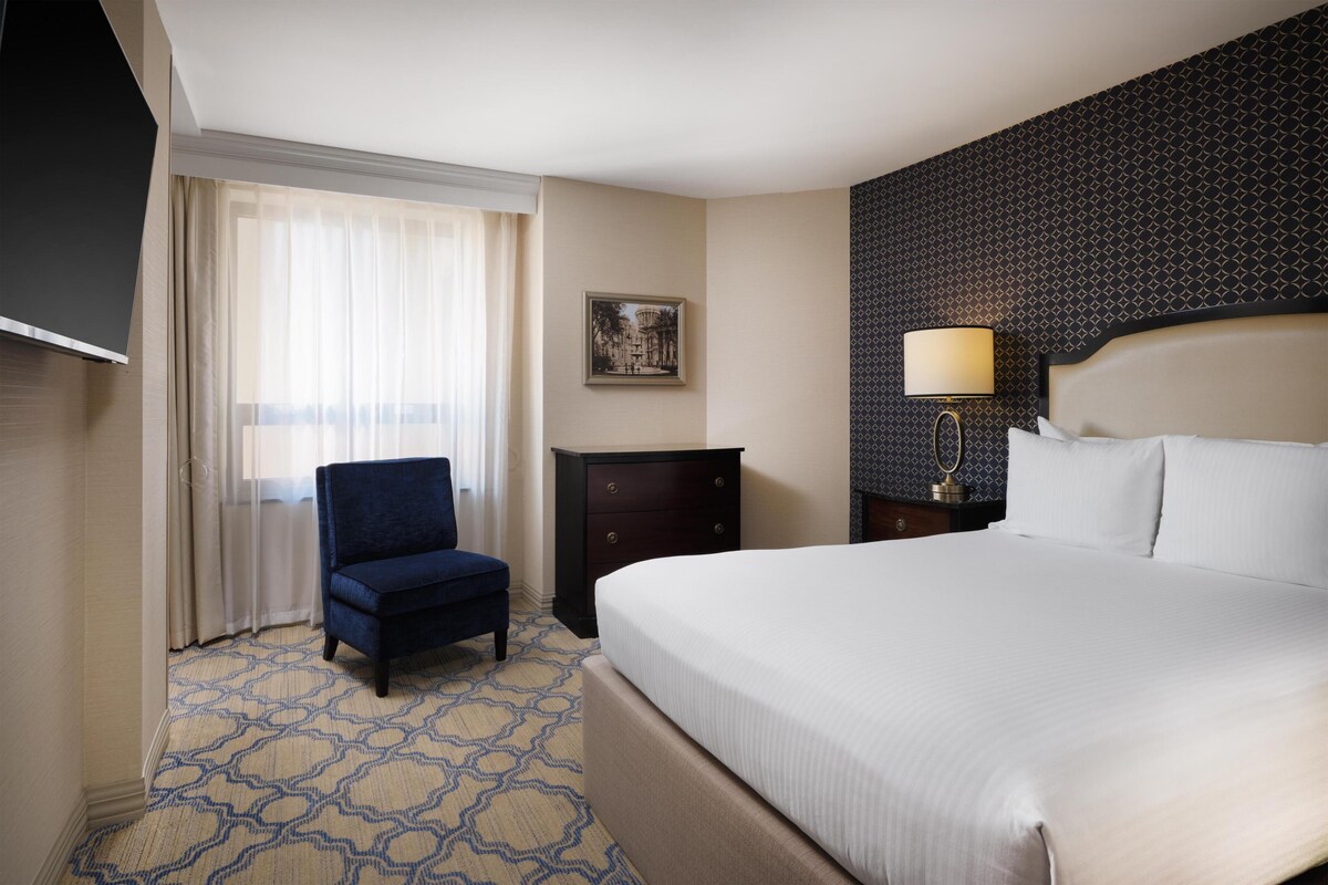 A hotel room with a bed, a chair, a dresser, a television, and a window with curtains.