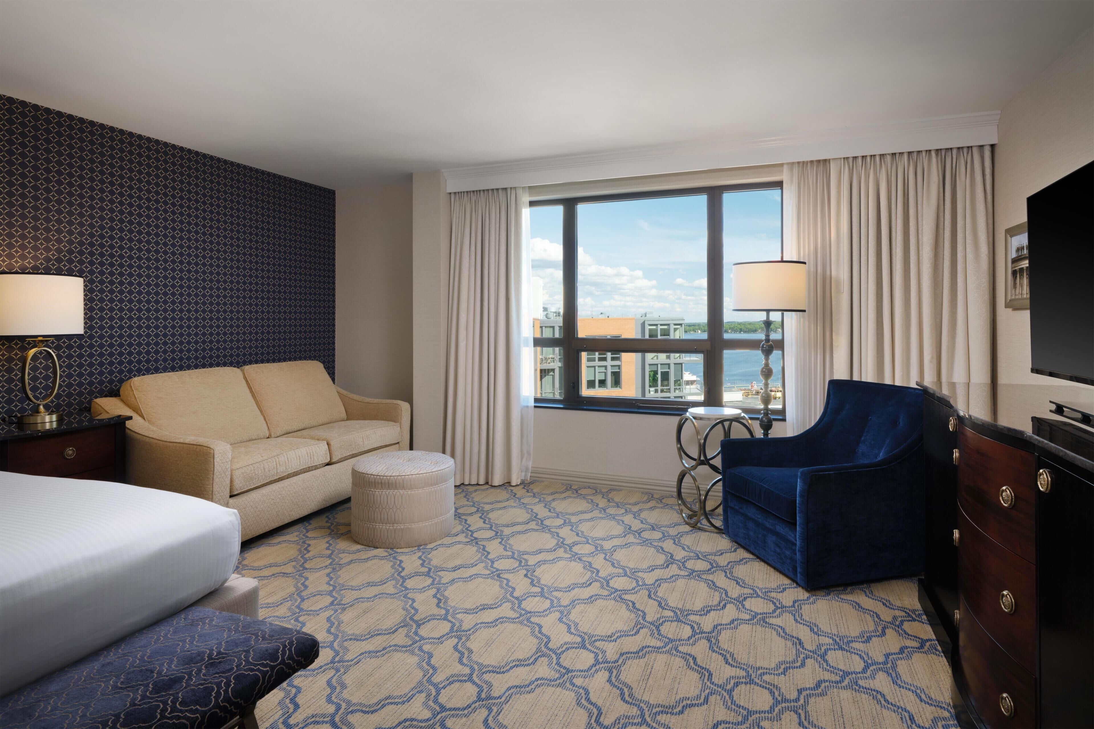 Habitación de hotel con cama, sofá, sillón, una pequeña otomana, escritorio con lámpara, televisión y ventana grande con cortinas.