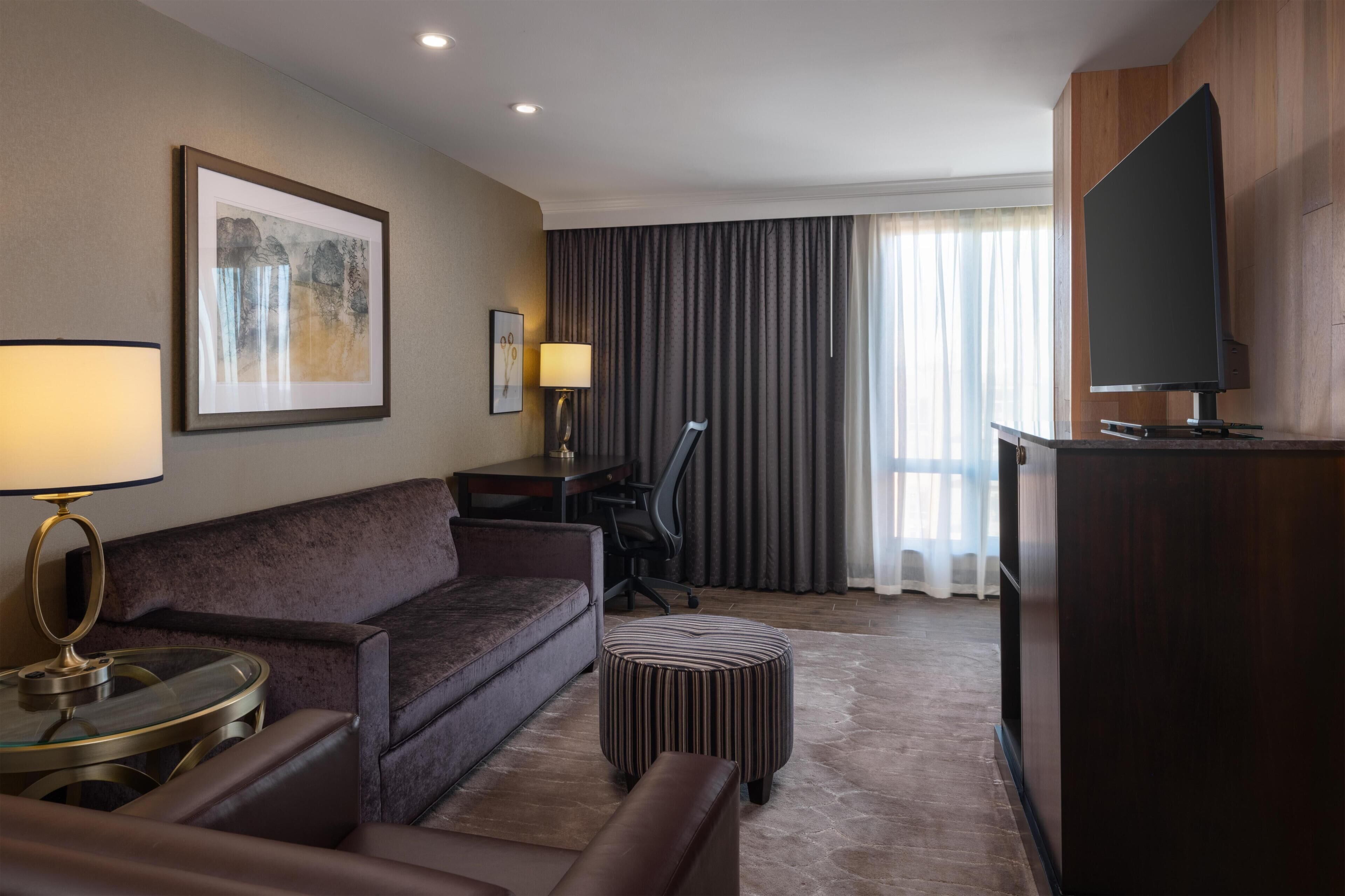 Ein Hotelzimmer mit Sofa, Schreibtisch, Fernseher, Lampe und einem Fenster mit Vorhängen.